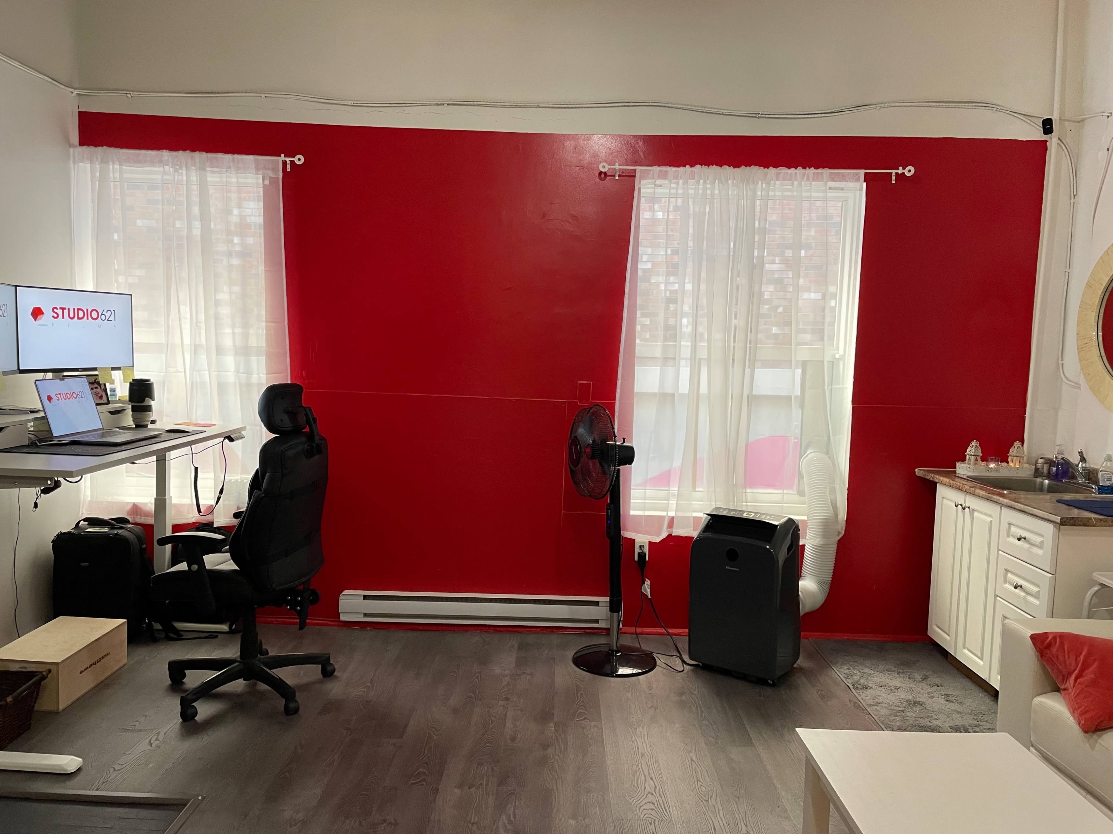 A room with a red accent wall, a workspace featuring a desk and computer, a black office chair, a fan, and a kitchenette area.