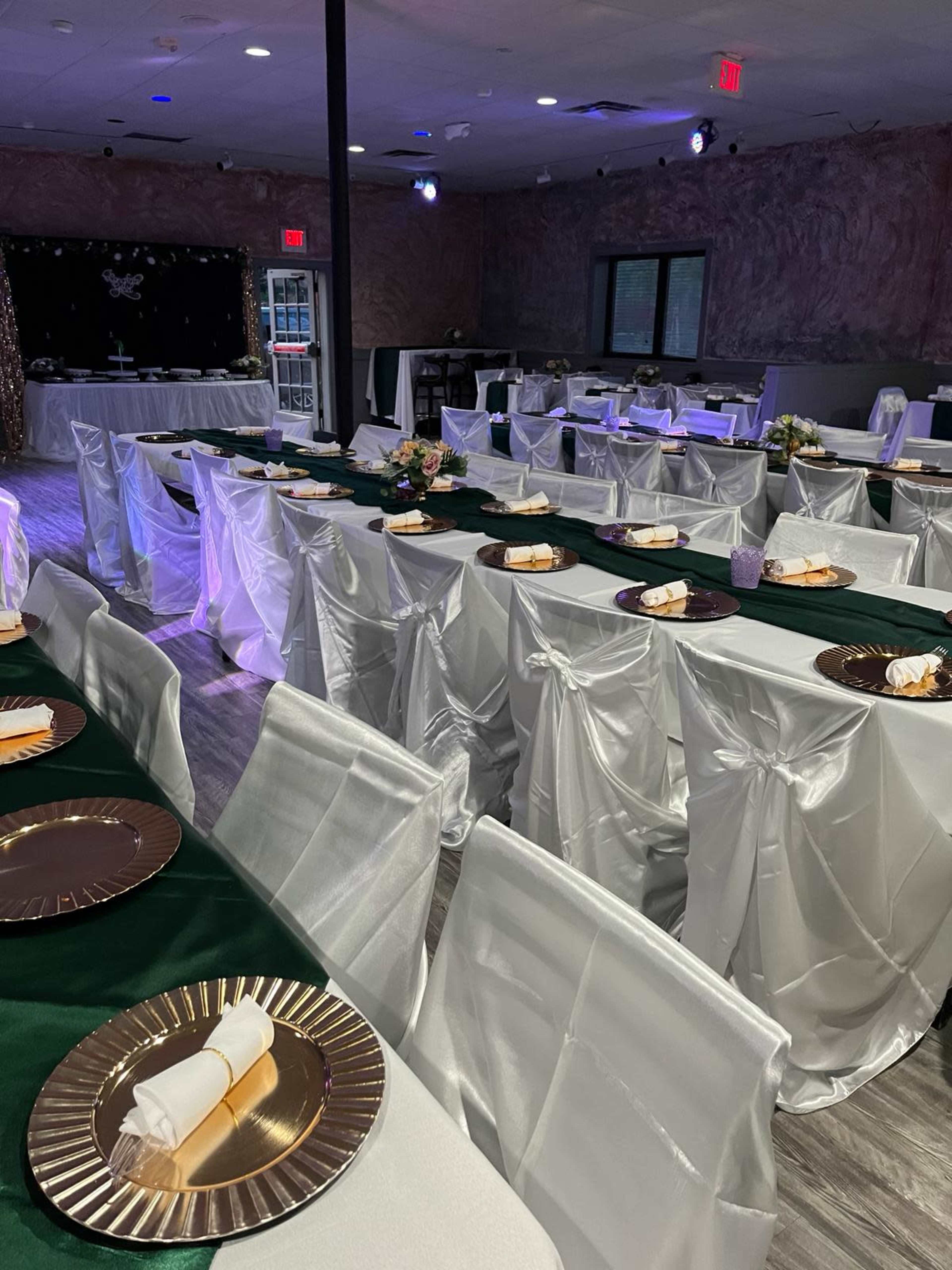A banquet hall set up with tables dressed in green and white tablecloths, adorned with gold-rimmed plates and neatly arranged napkins.
