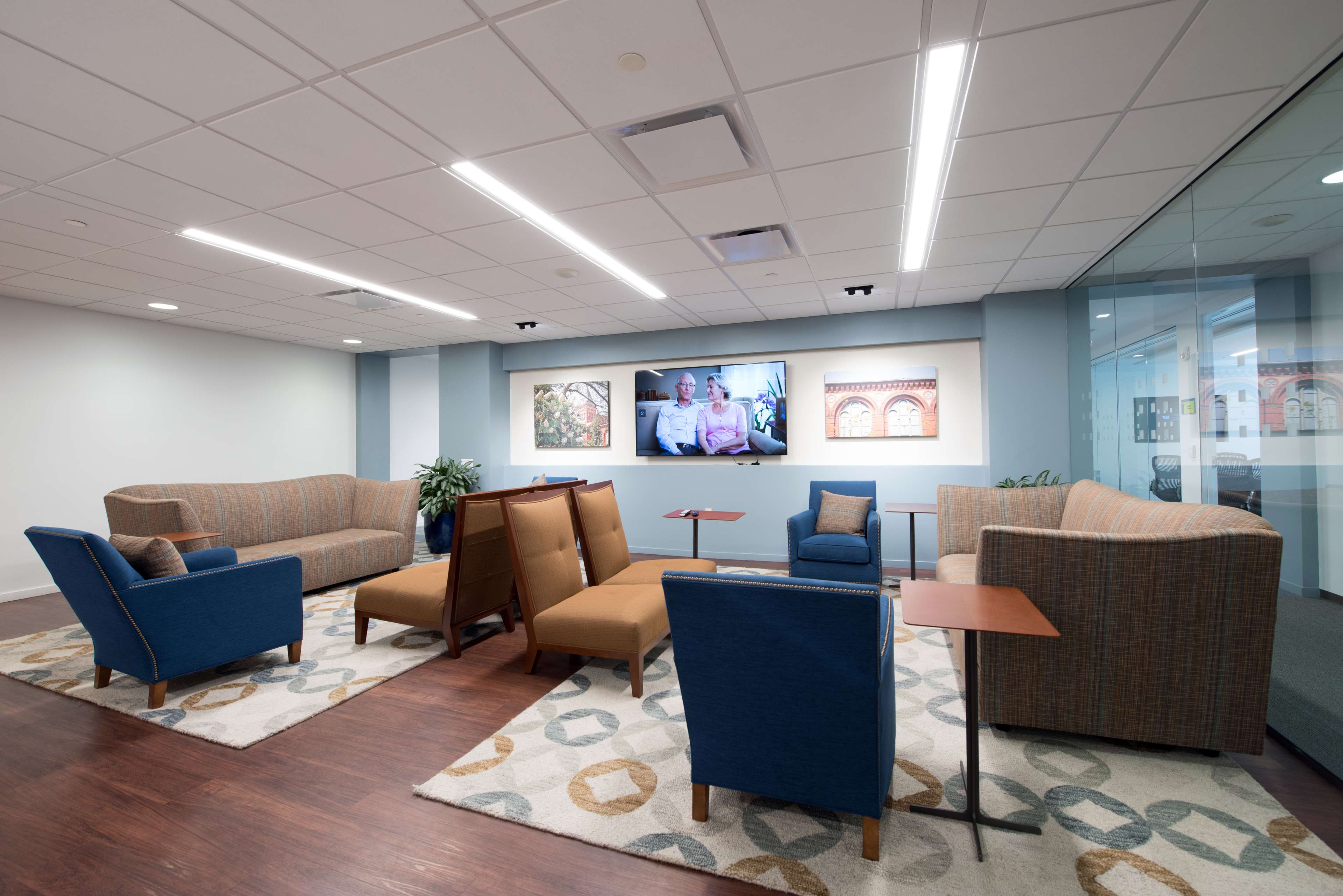 A modern lounge area features various seating arrangements around a central television, with patterned rugs on the floor.