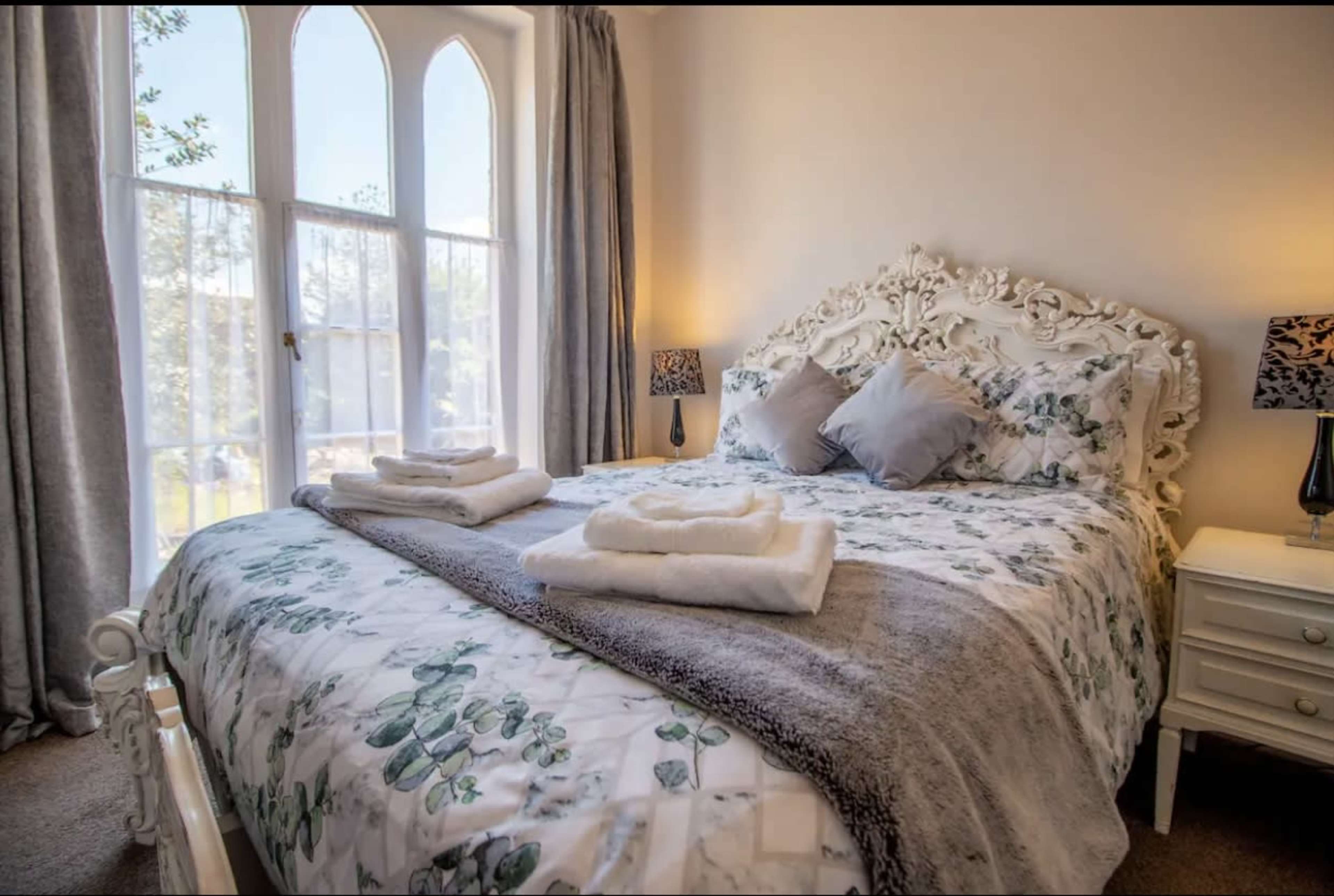 A neatly arranged bedroom features a floral-patterned bedspread with white towels folded on the bed, flanked by two lamps and large windows.