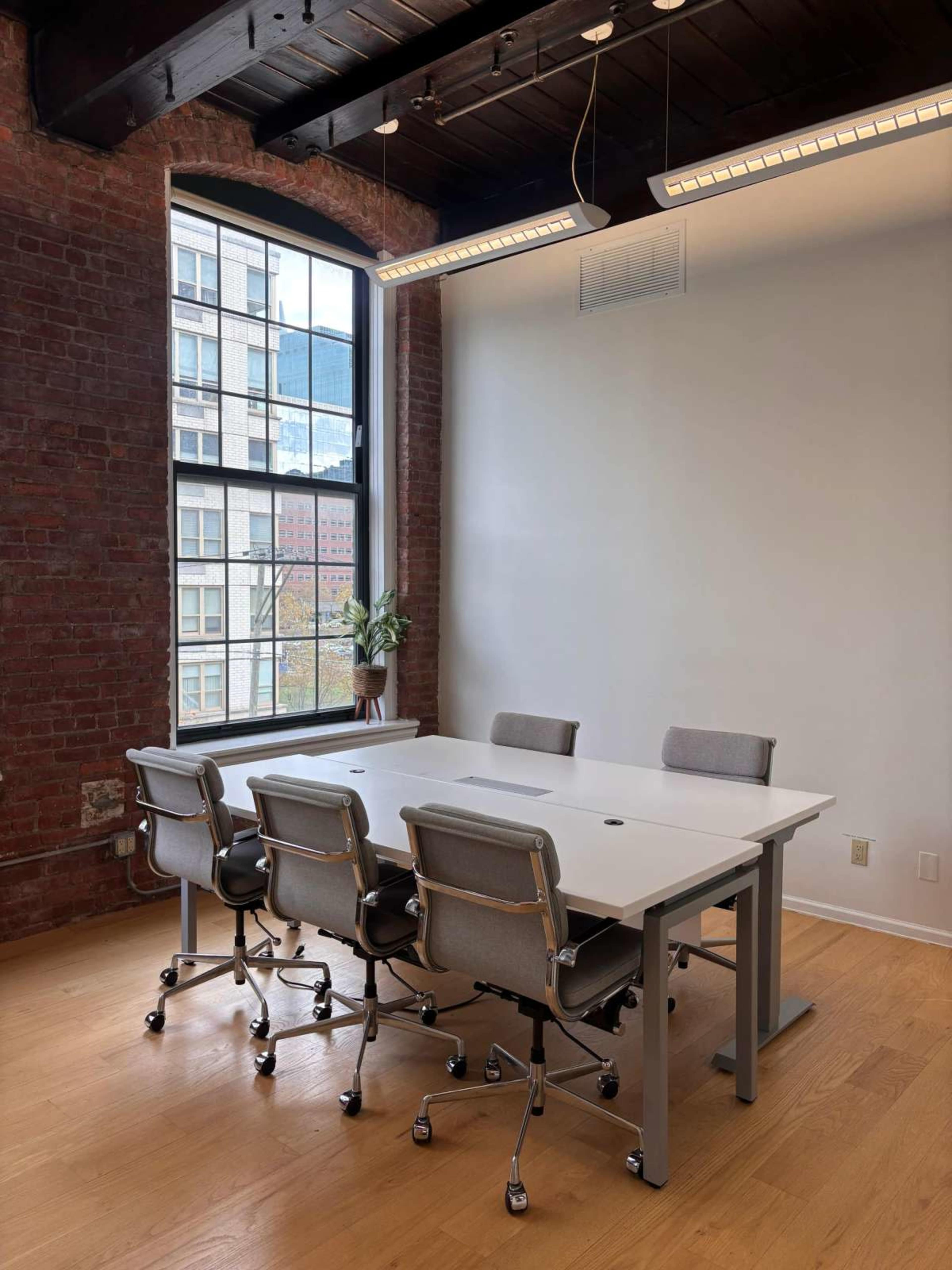 A modern conference room features a long white table surrounded by four ergonomic chairs, with a large window revealing a view of a cityscape.