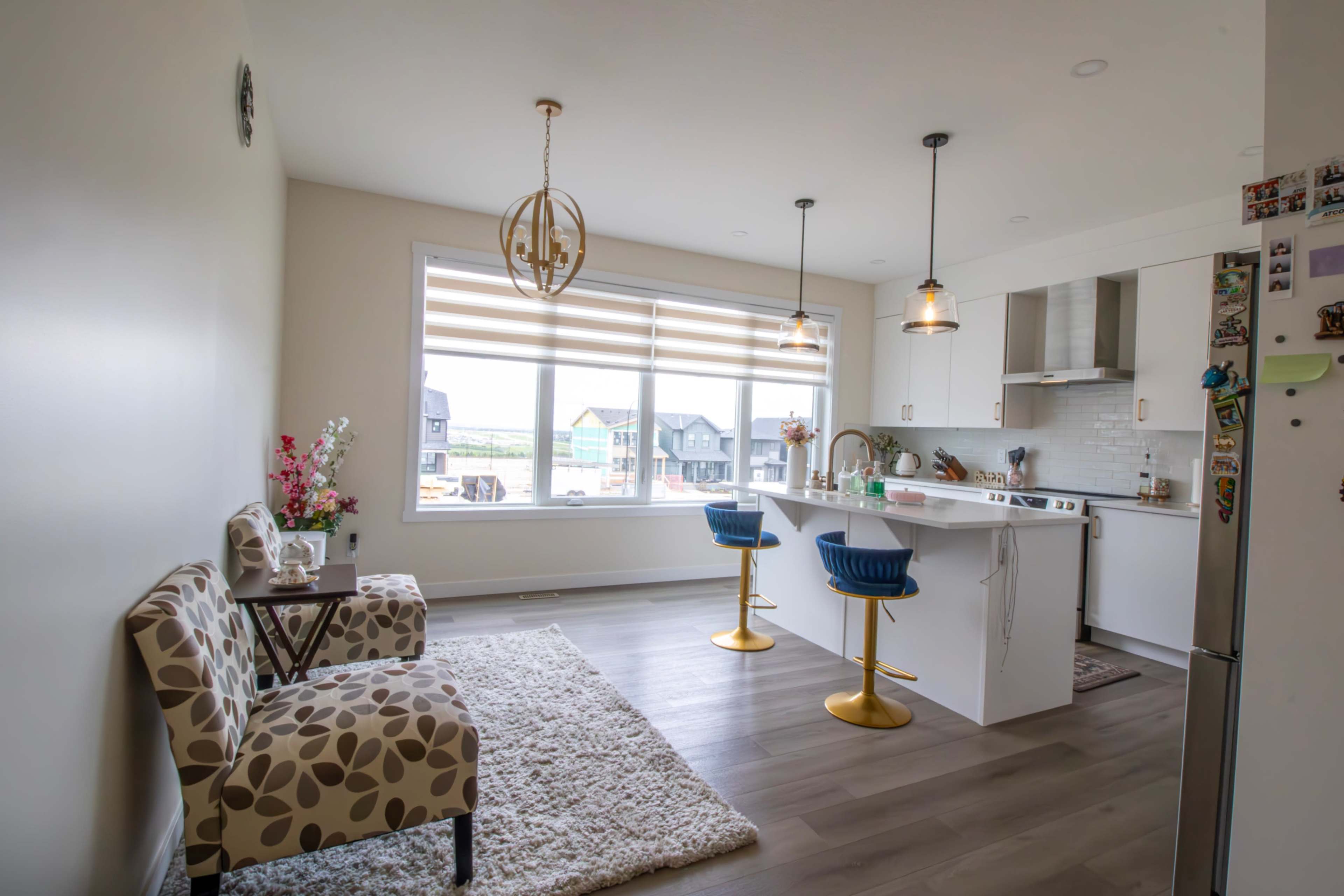 The image shows a modern kitchen and dining area with a covered island, two blue bar stools, a small seating area, and large windows providing natural light.