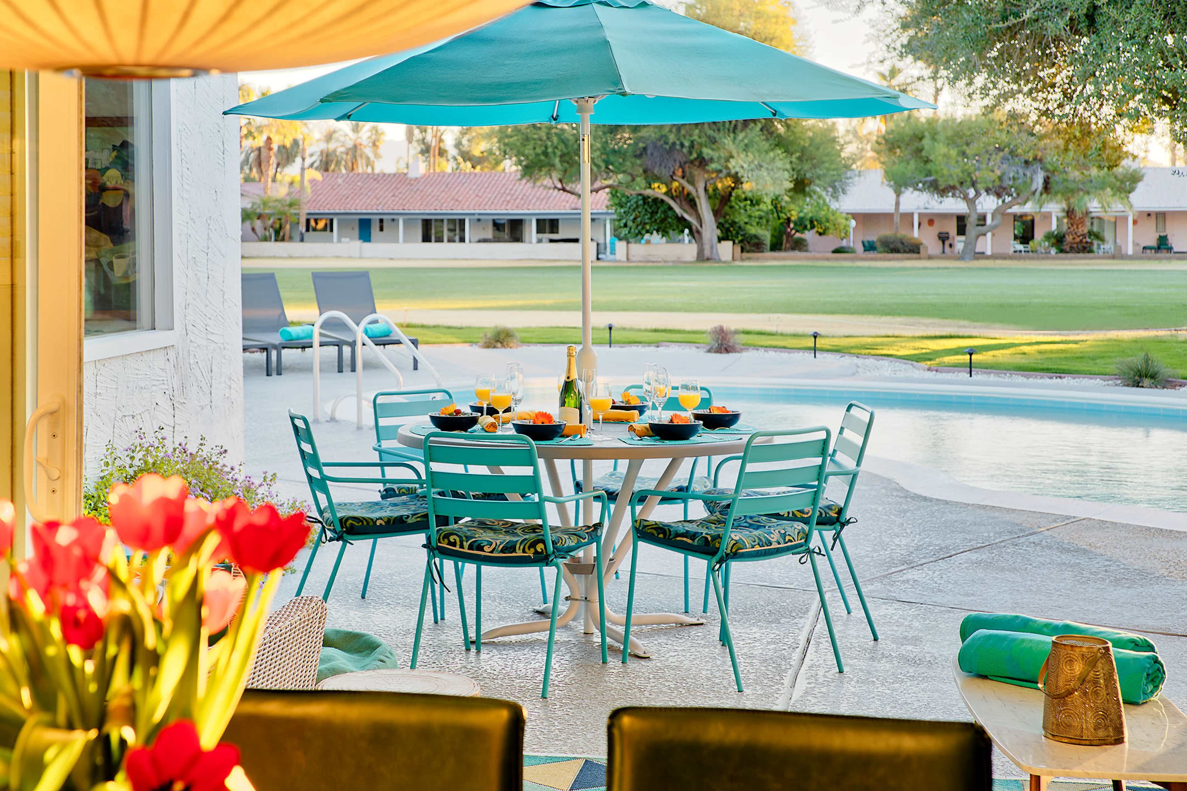 A patio area features a round table with teal chairs set for brunch, shaded by a large umbrella, beside a swimming pool and overlooking a grassy landscape.
