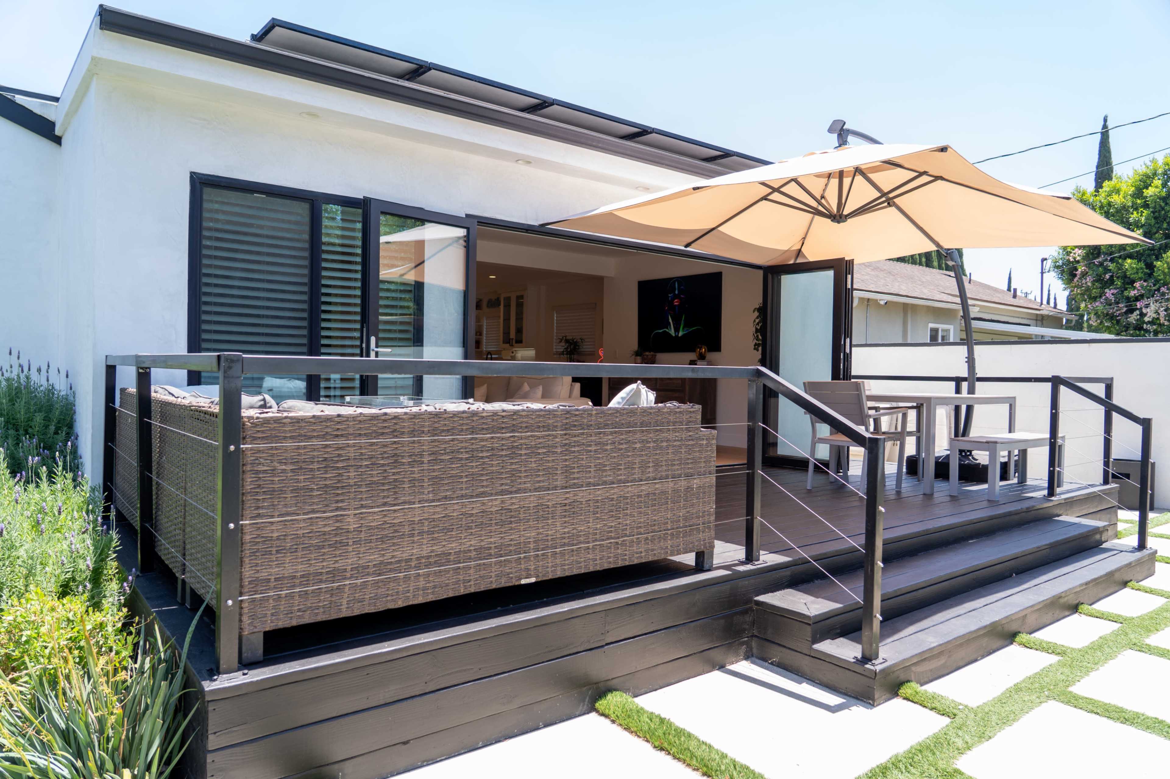 The image shows a modern patio area with a wicker sofa, an umbrella for shade, and steps leading up to an indoor living space.