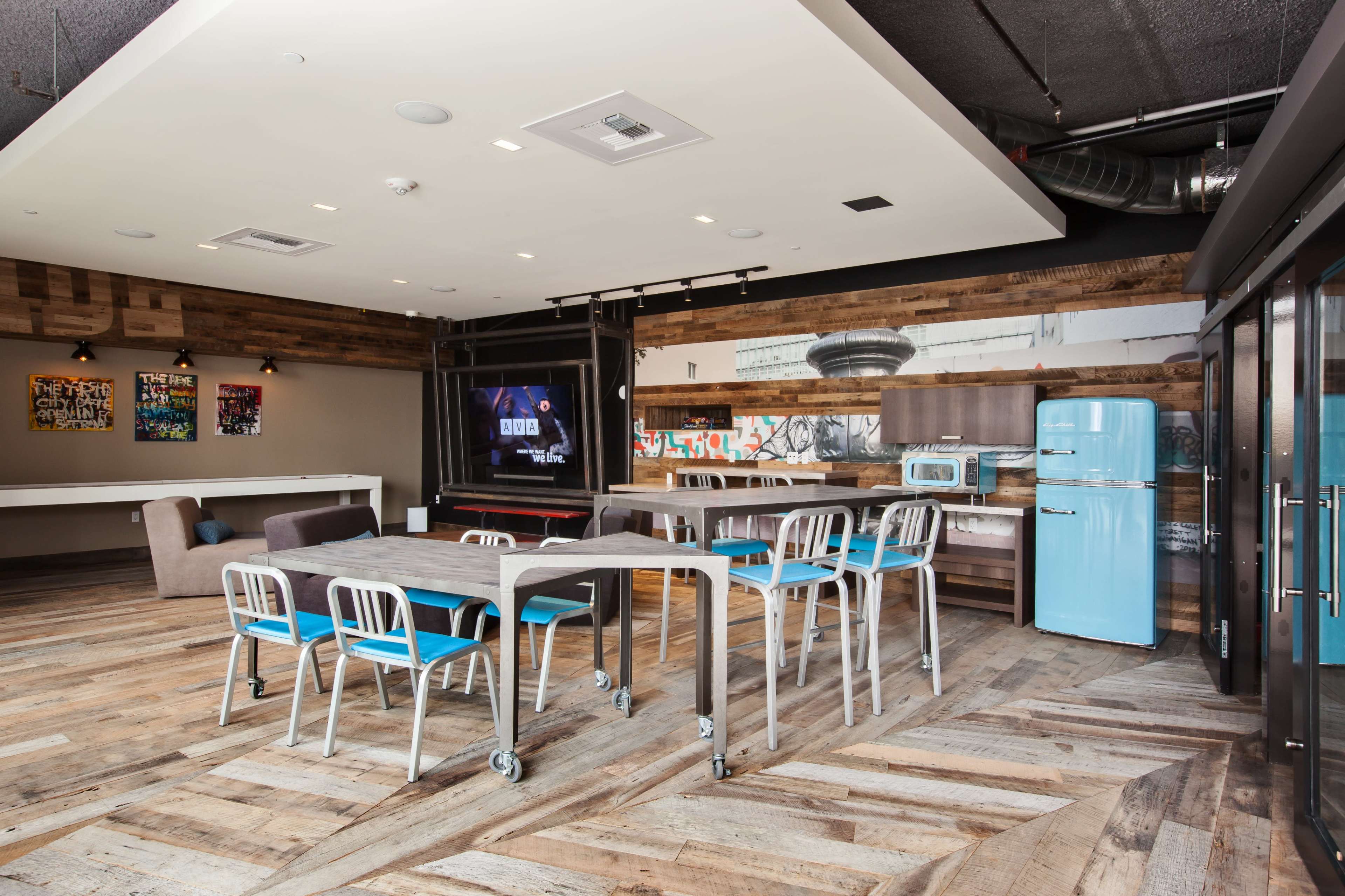 The image shows a modern office space featuring a large dining table surrounded by metal chairs, a retro blue refrigerator, and wooden accents on the walls and floor.
