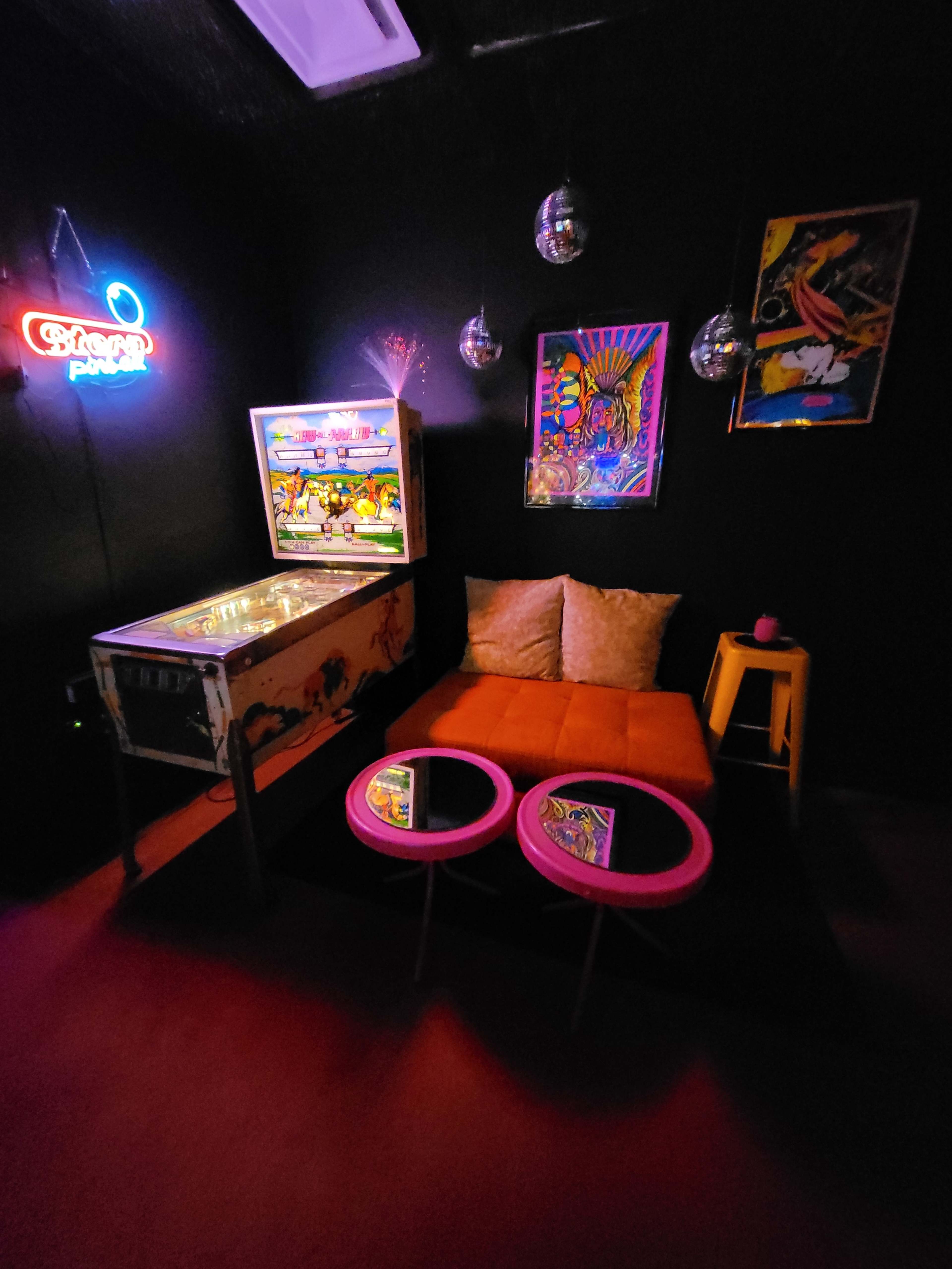 A cozy lounge area with an orange couch, two pink coffee tables, a vintage pinball machine, and colorful artwork on a black wall.