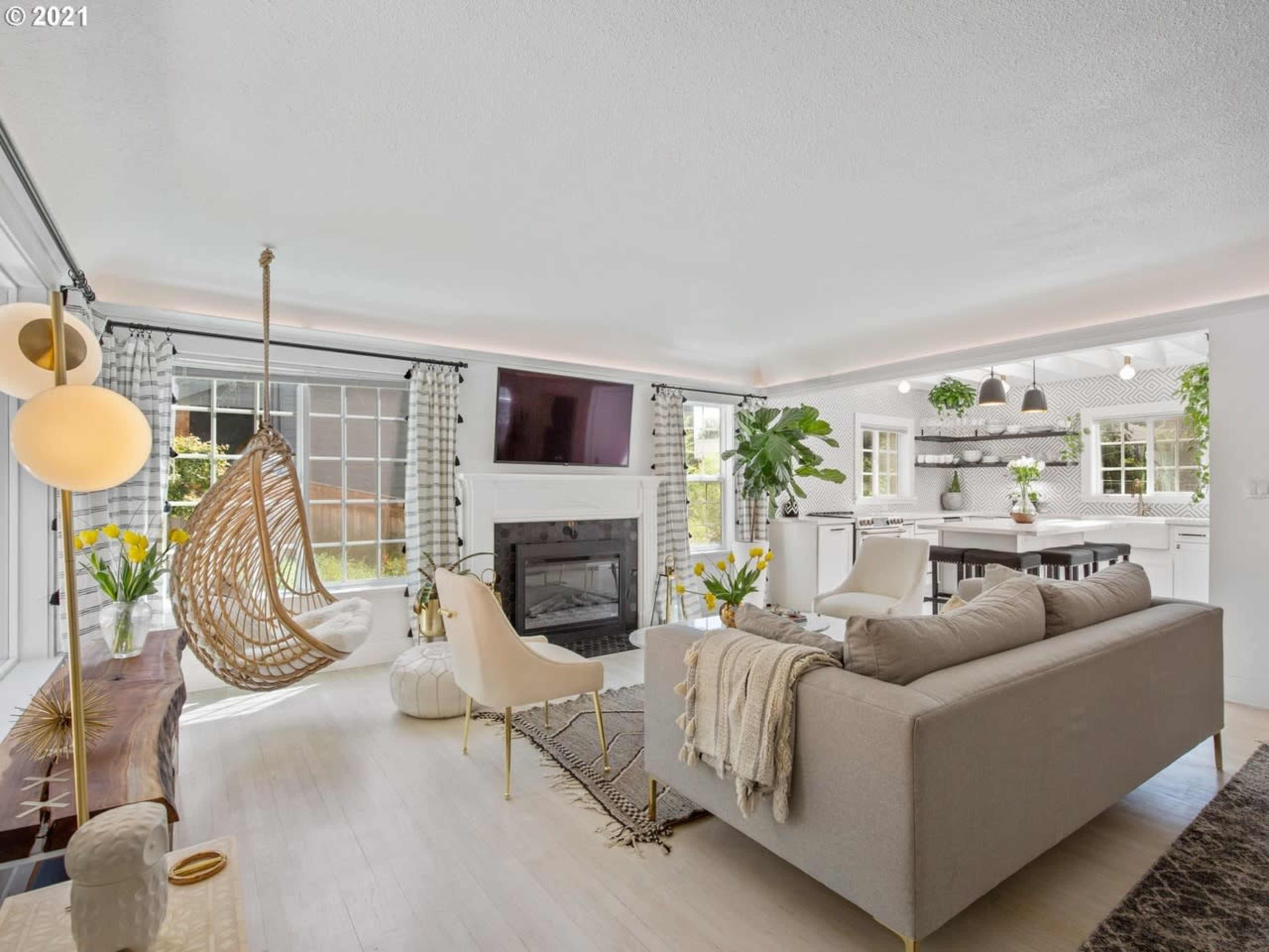 A modern living room features a hanging chair, a fireplace, large windows, and an open layout leading to a kitchen area.