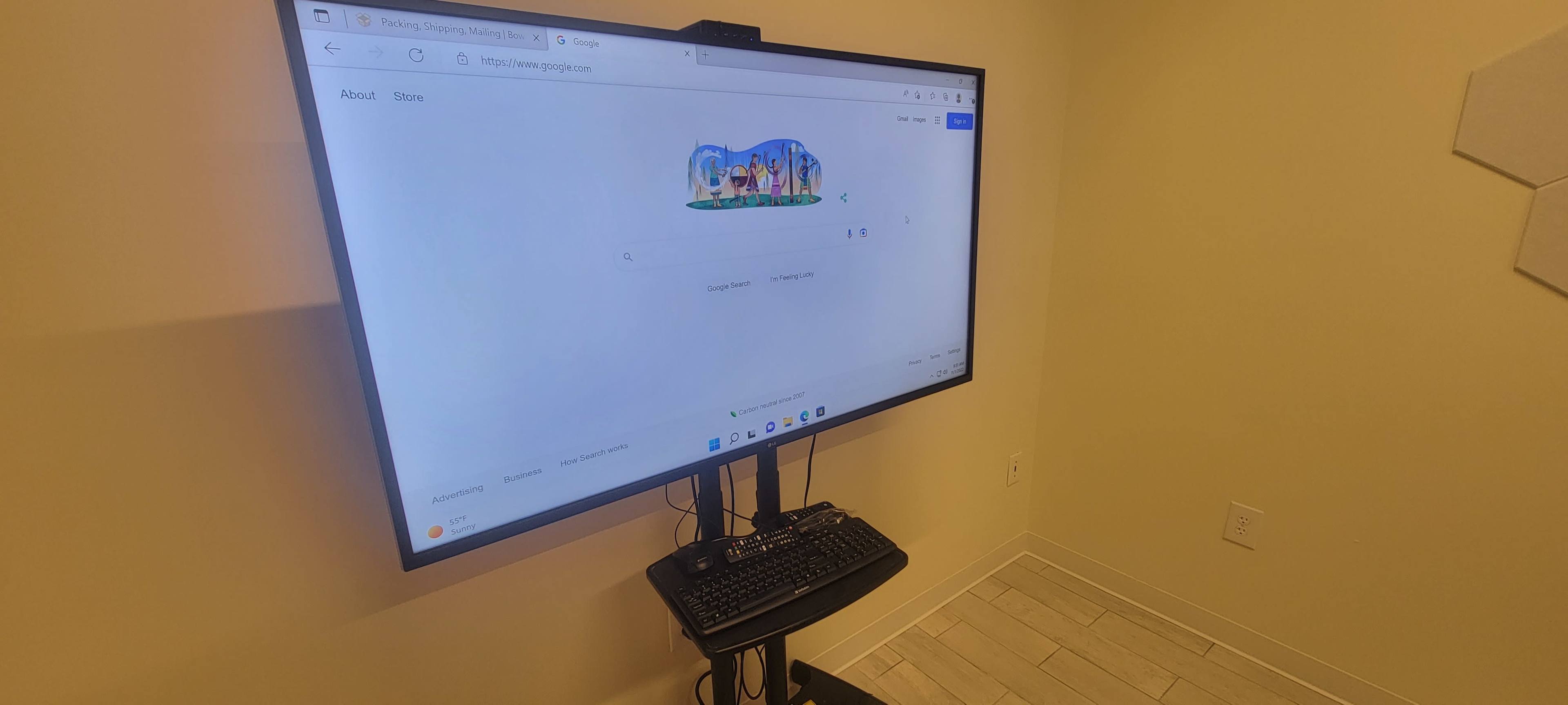 A large monitor displaying the Google search homepage is mounted on a stand in a room with light-colored walls and flooring.