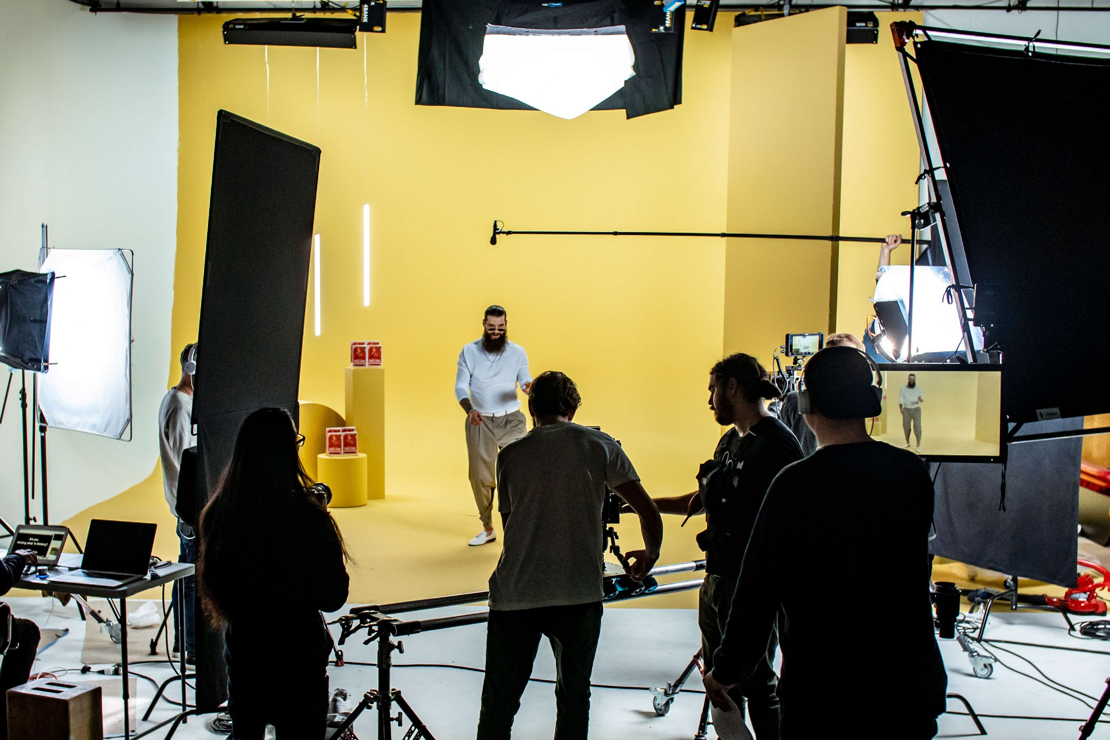 A film crew is set up in a studio with yellow walls, capturing a scene featuring a man in a white outfit.