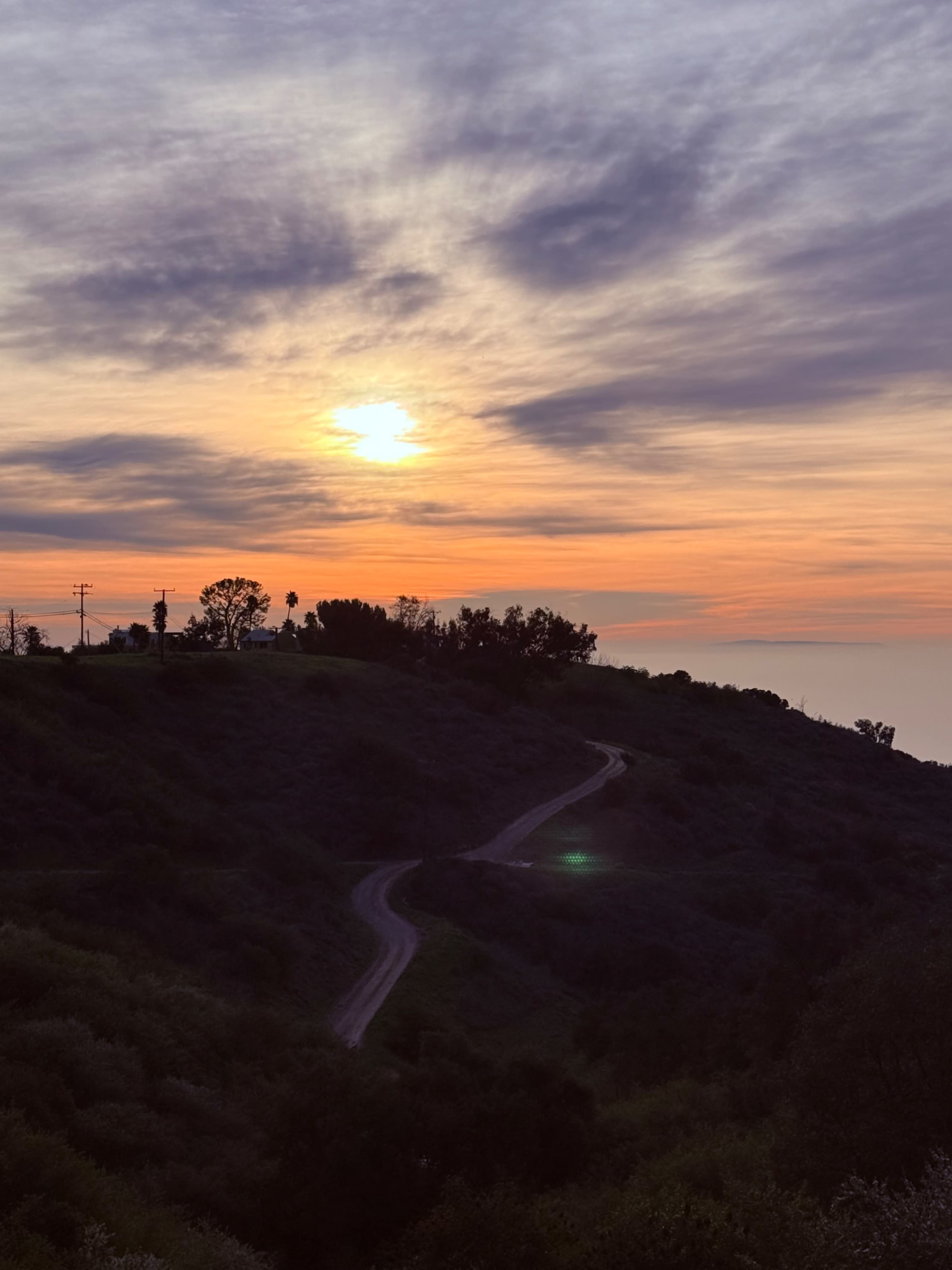 The image shows a winding dirt road descending through green hills under a sunset sky filled with clouds.