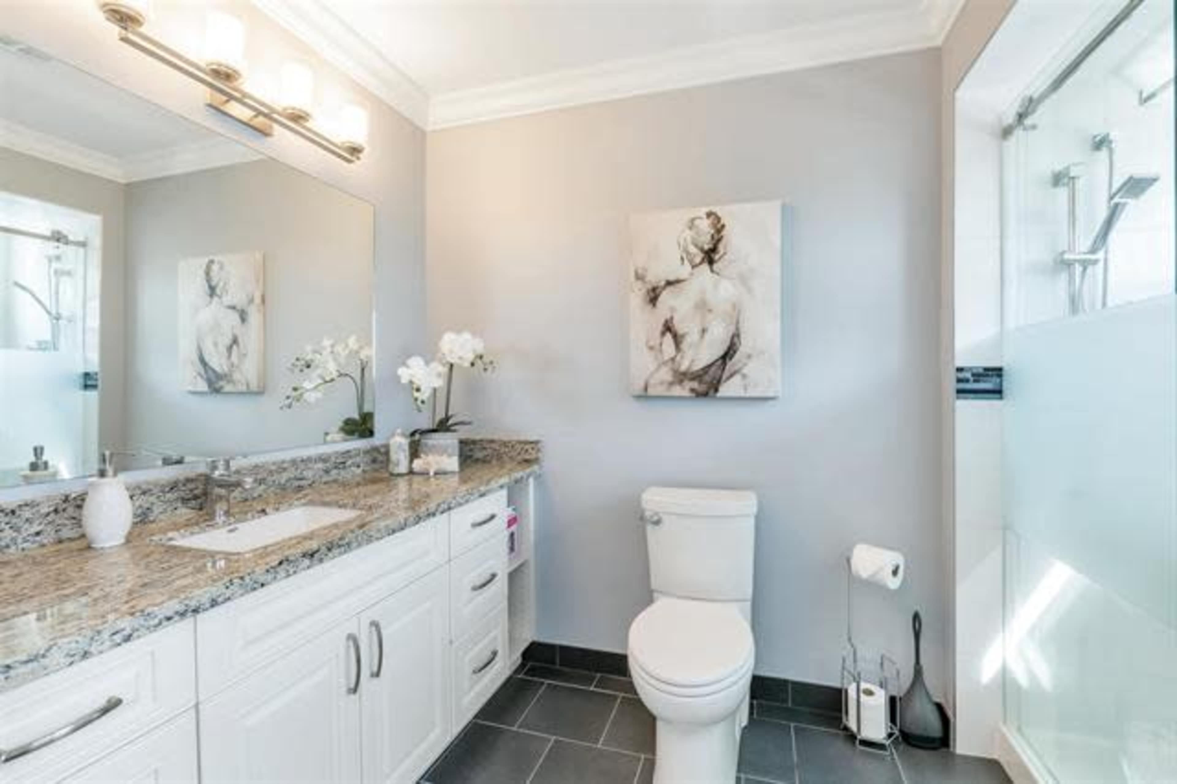 A modern bathroom featuring a light gray color scheme, a granite countertop with a double sink, a white toilet, and a large mirror above the sink.