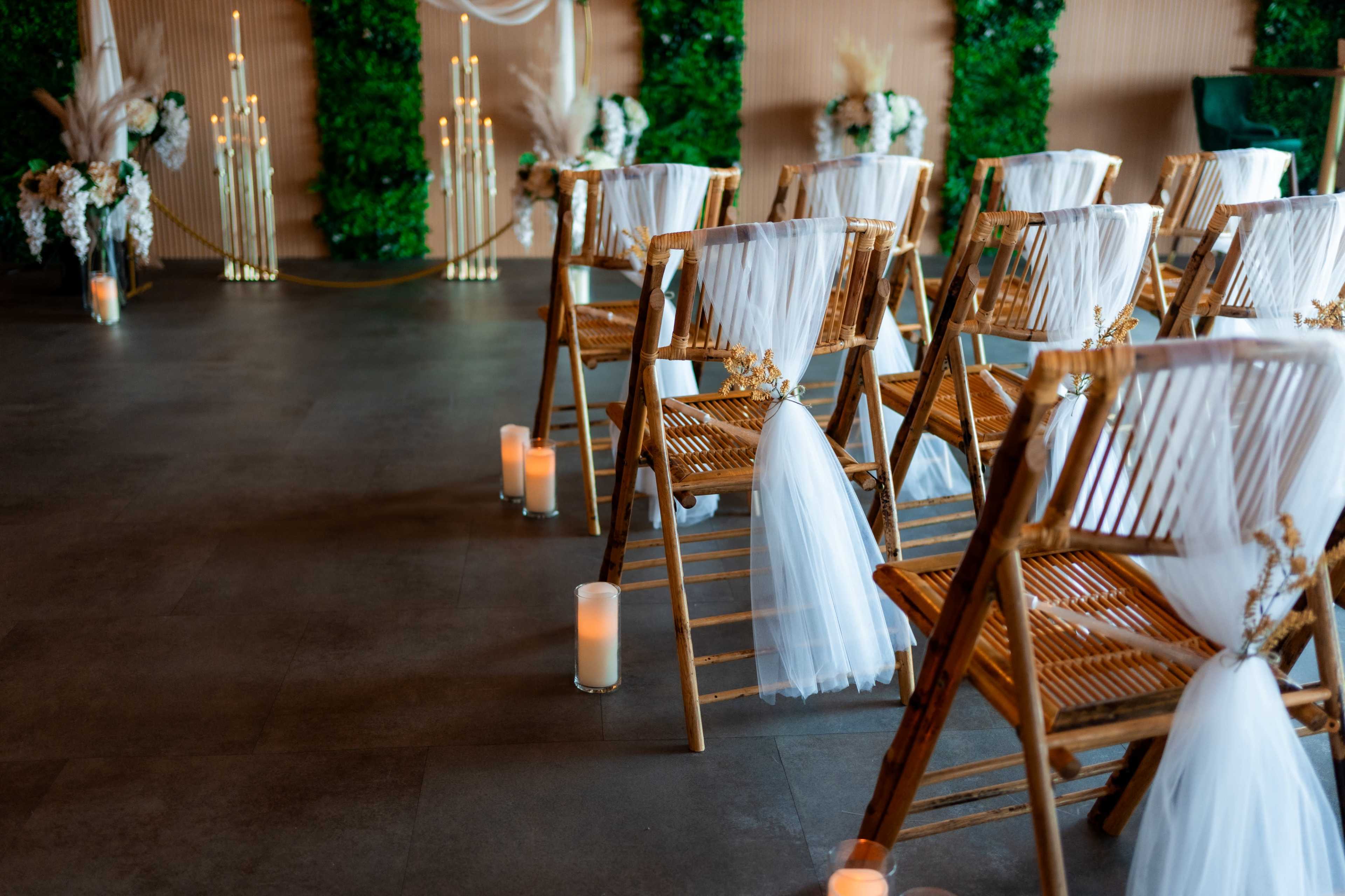 The image shows a set of bamboo chairs with white decorations arranged in an indoor wedding venue, surrounded by greenery and lit candles.