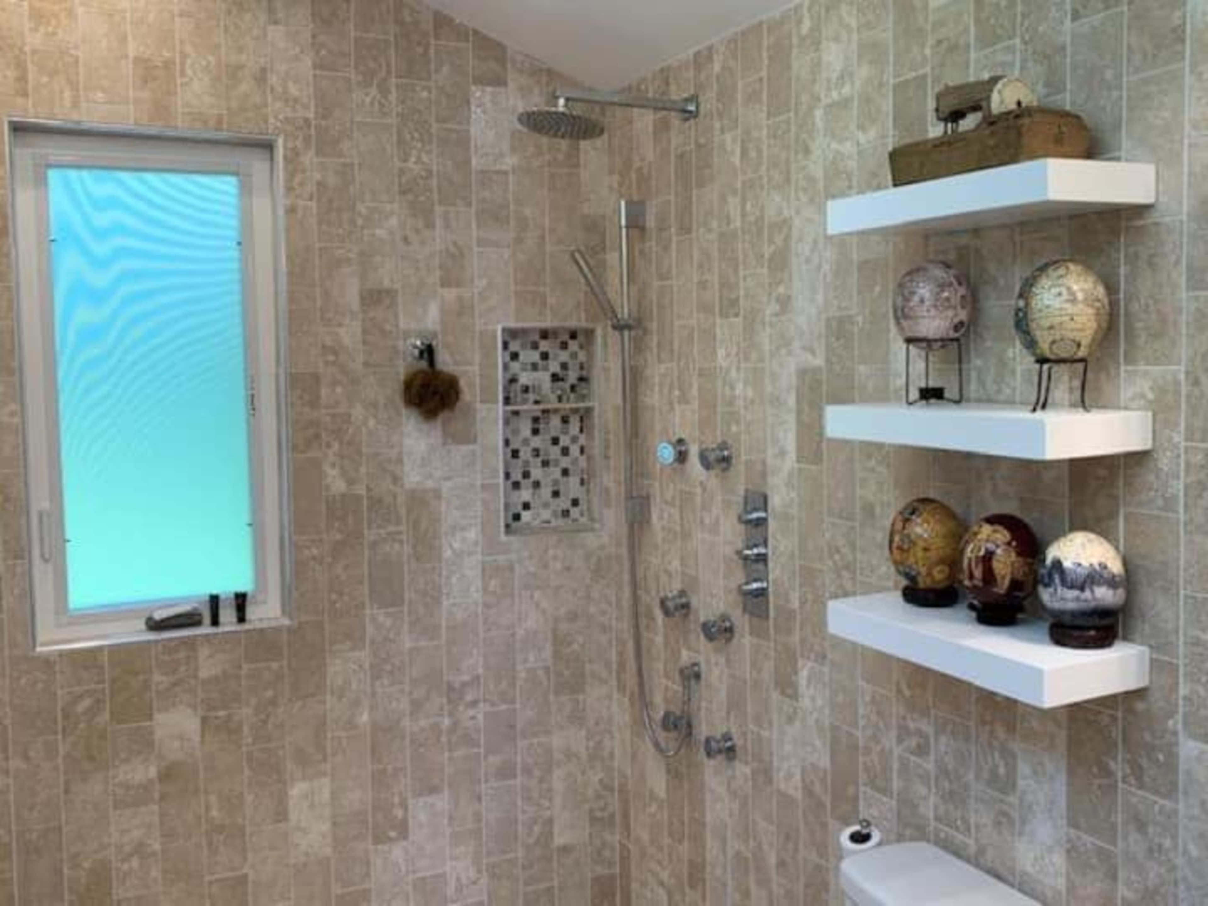 The image shows a modern bathroom shower area with stone tile walls, a glass window, and decorative shelves displaying various ornamental globes.