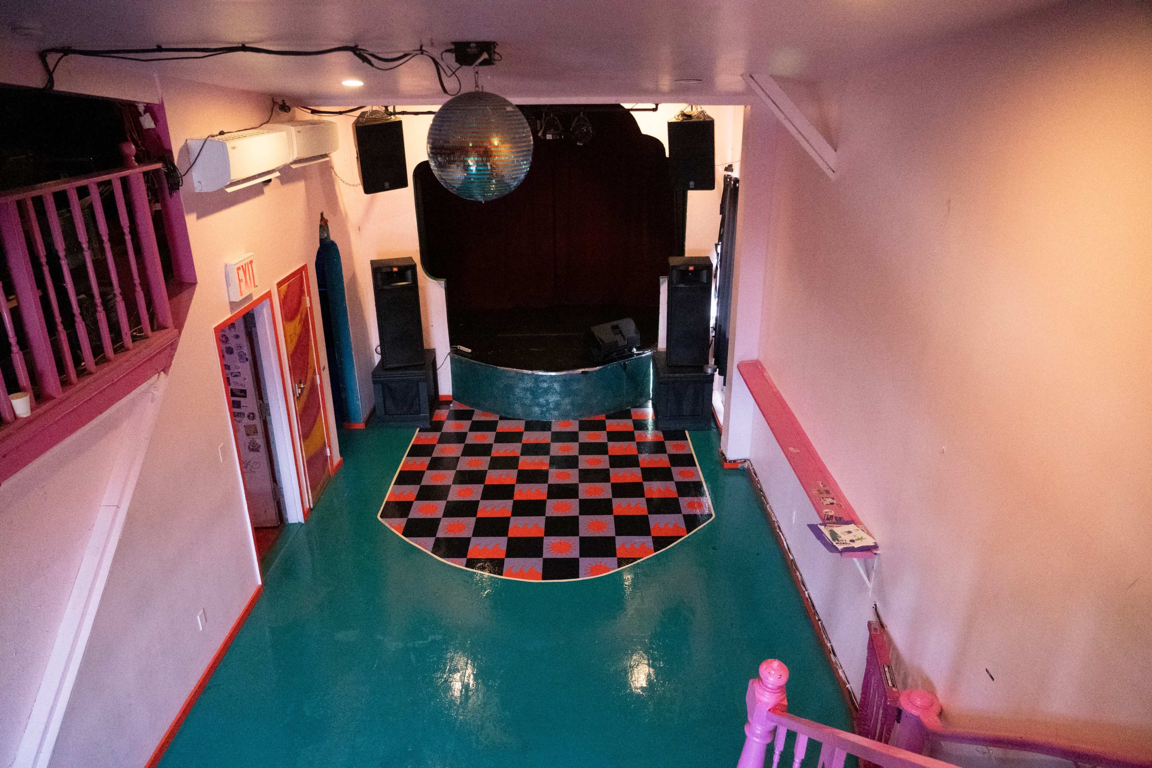 A colorful music venue with a checkered dance floor, a small stage, and pink-painted walls.