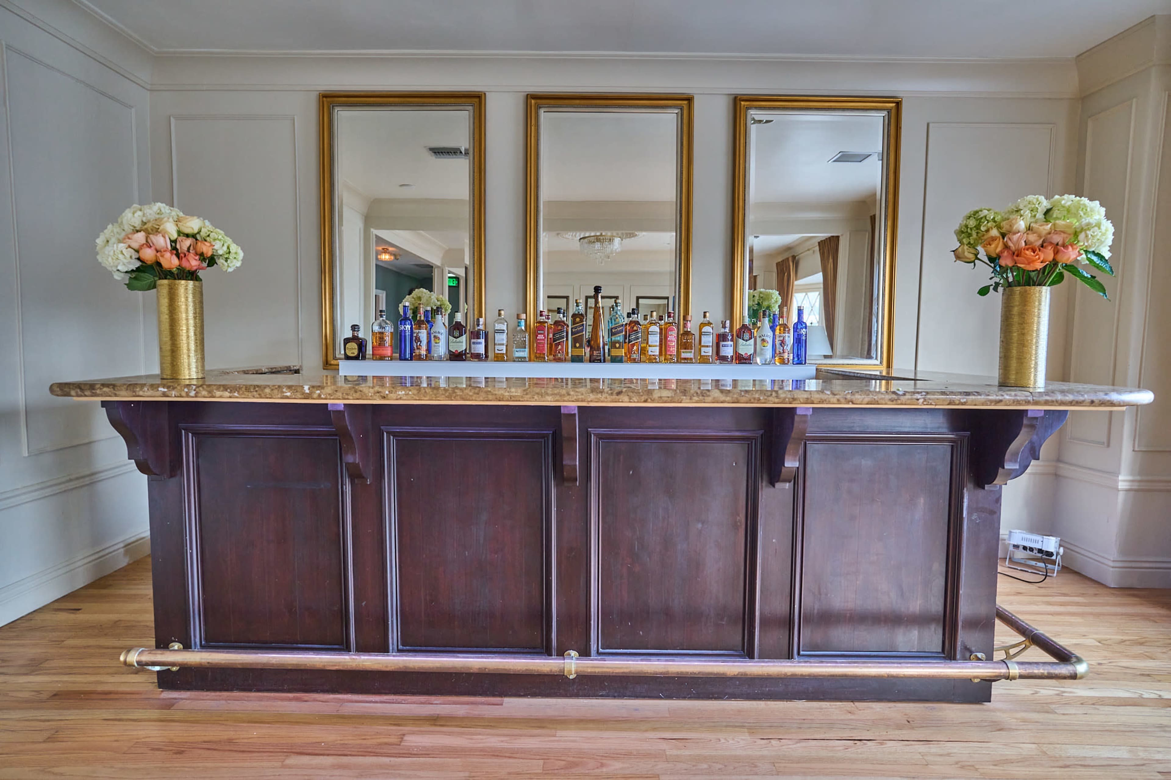 A wooden bar with a marble countertop displays a variety of colorful bottles and is flanked by three large mirrors and floral arrangements.