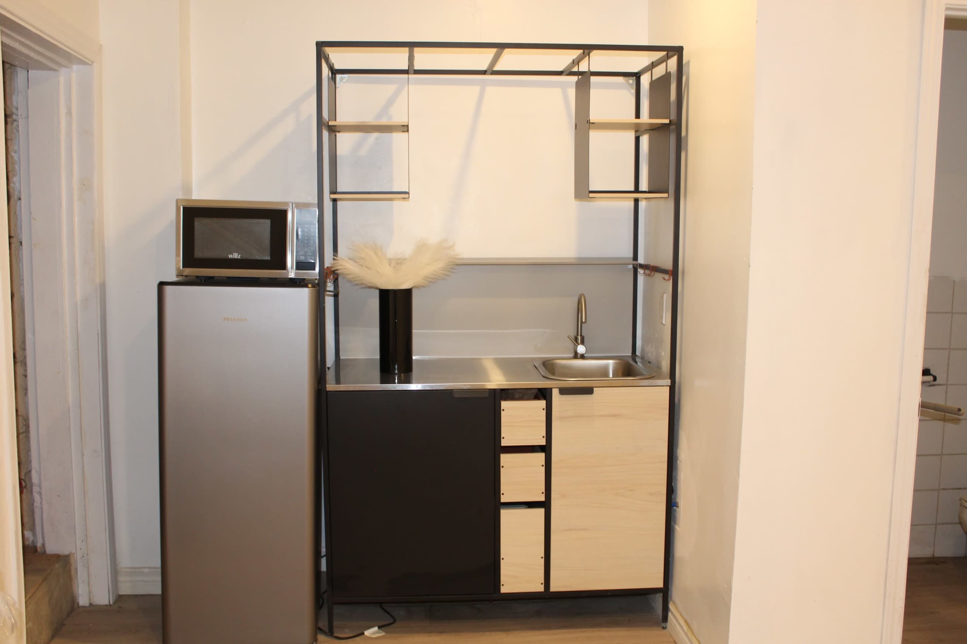 A modern kitchen setup featuring a stainless steel refrigerator, a microwave on a shelf, and a compact sink with cabinetry below.