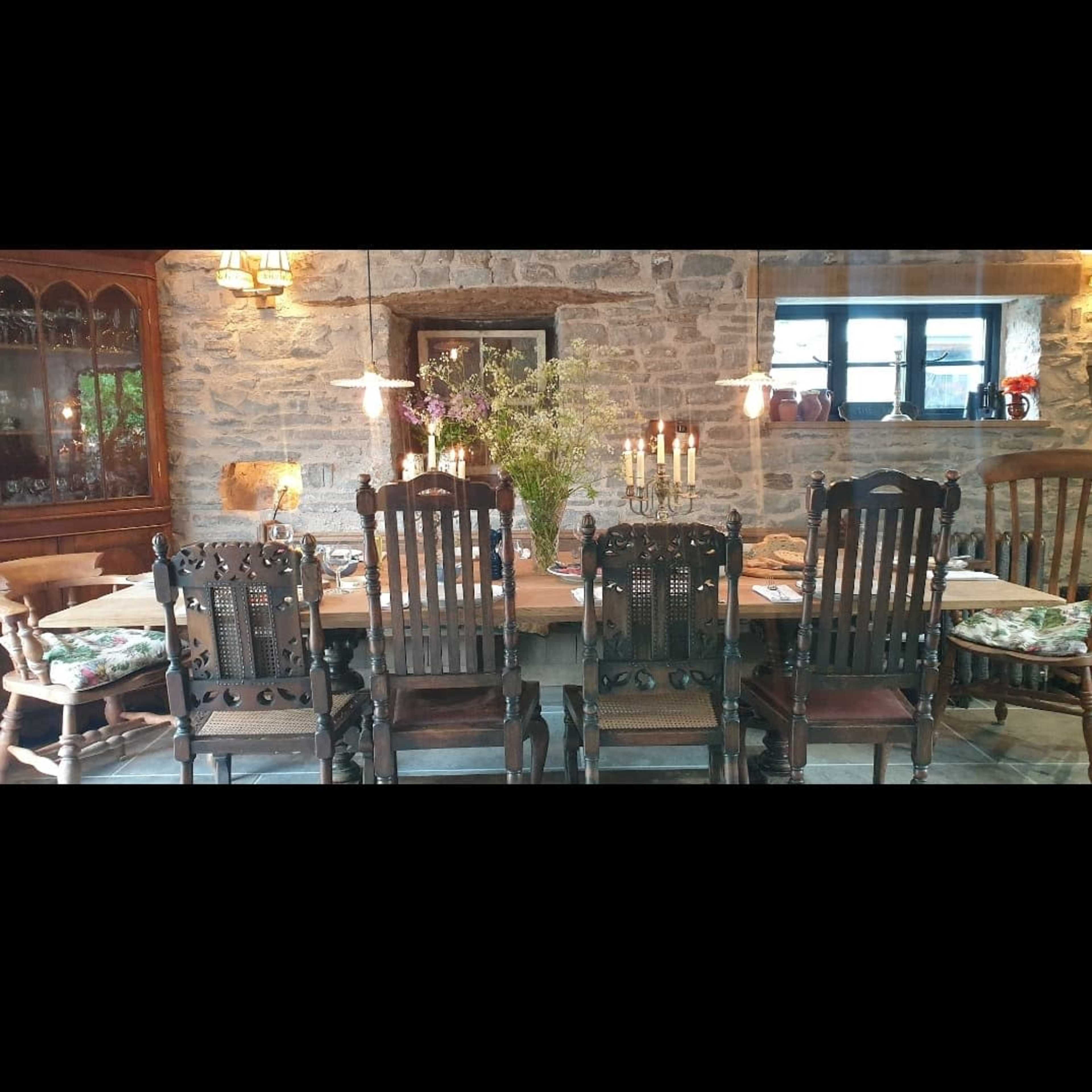 A rustic dining room features a long wooden table surrounded by high-backed chairs, with stone walls and warm lighting.