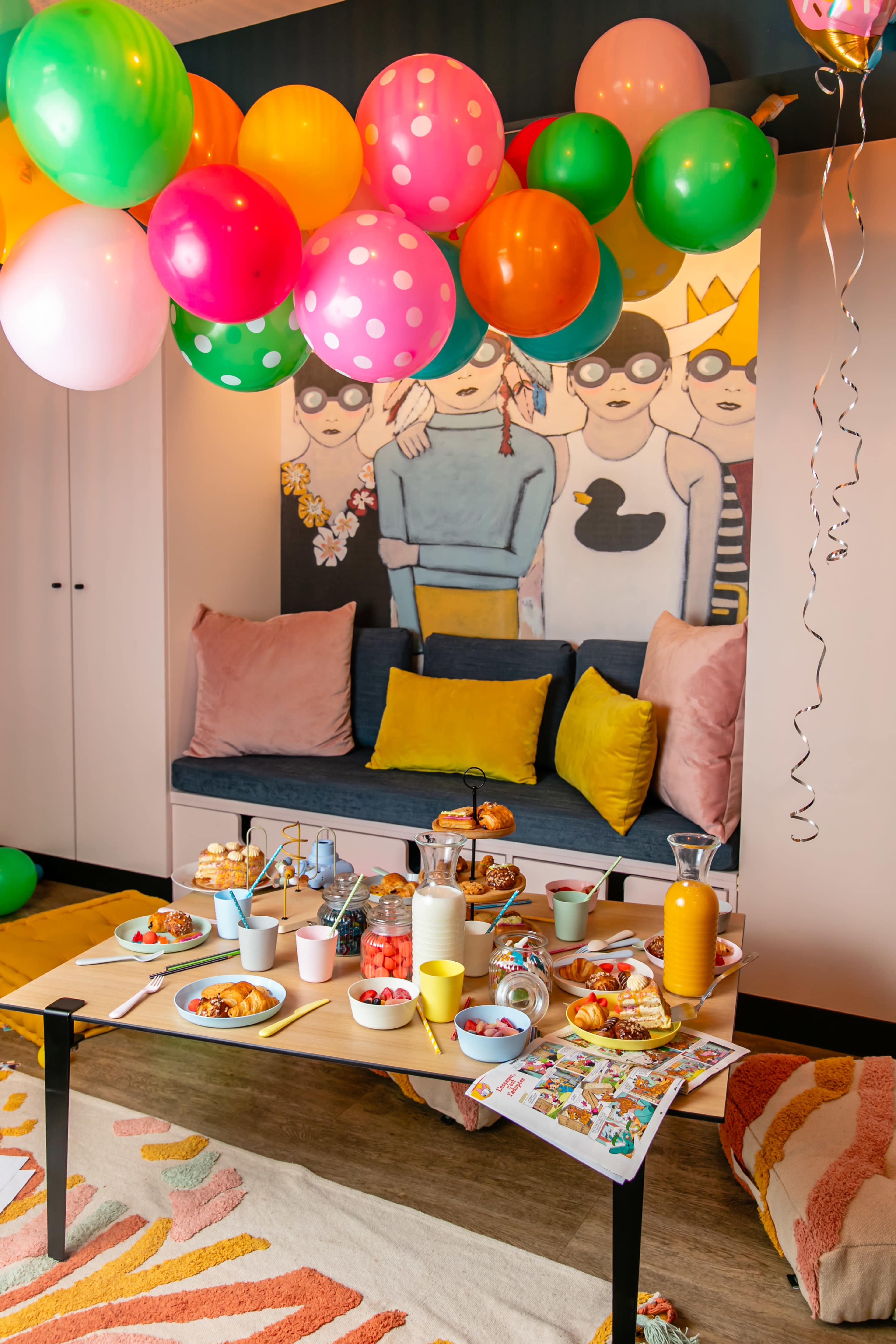 A colorful party setup features a table with various snacks and beverages, surrounded by balloons, while a mural of children adorns the wall behind a cozy seating area.