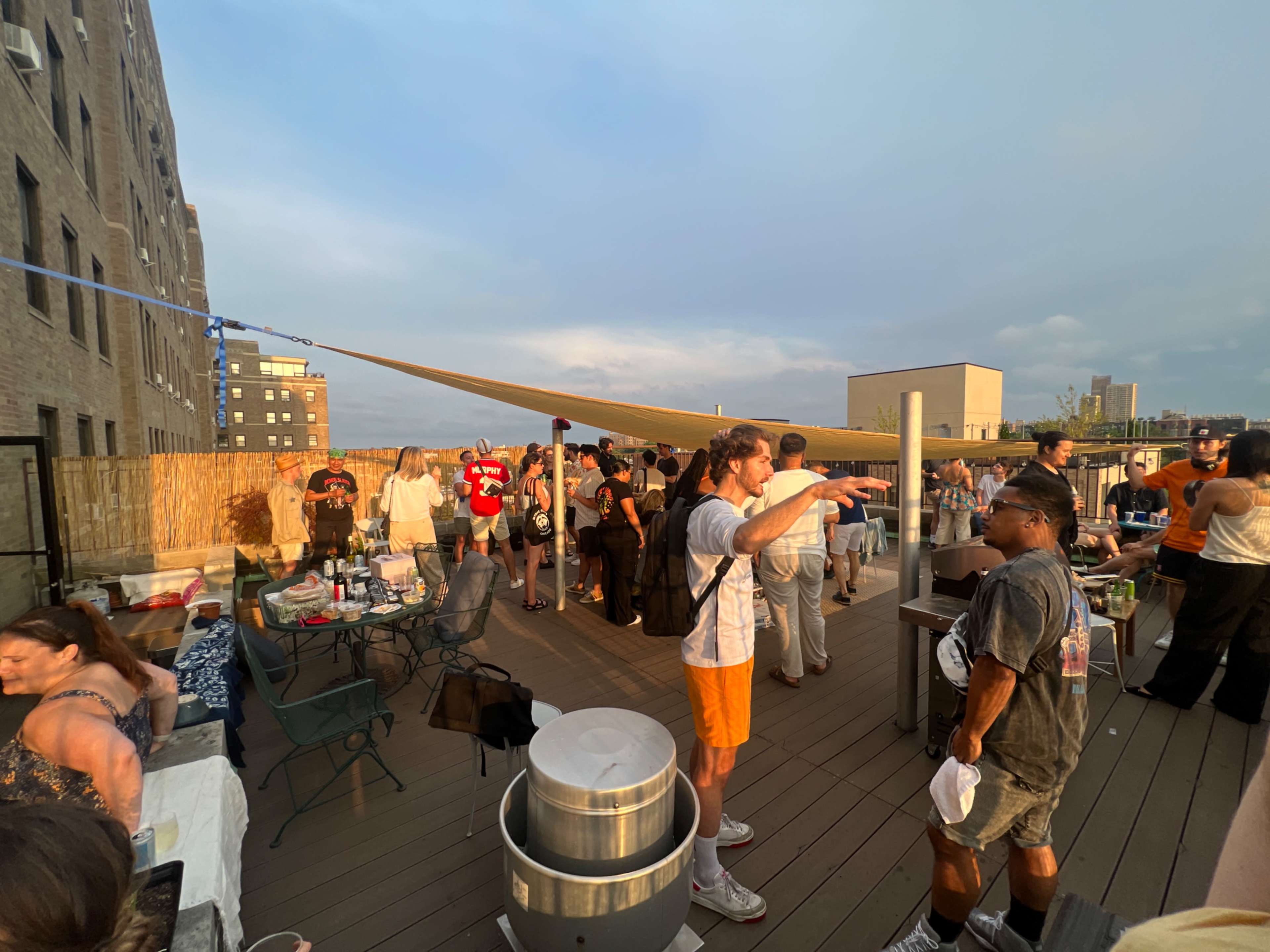 A crowd of people socializes at a rooftop gathering with tables, grills, and a large sunshade.