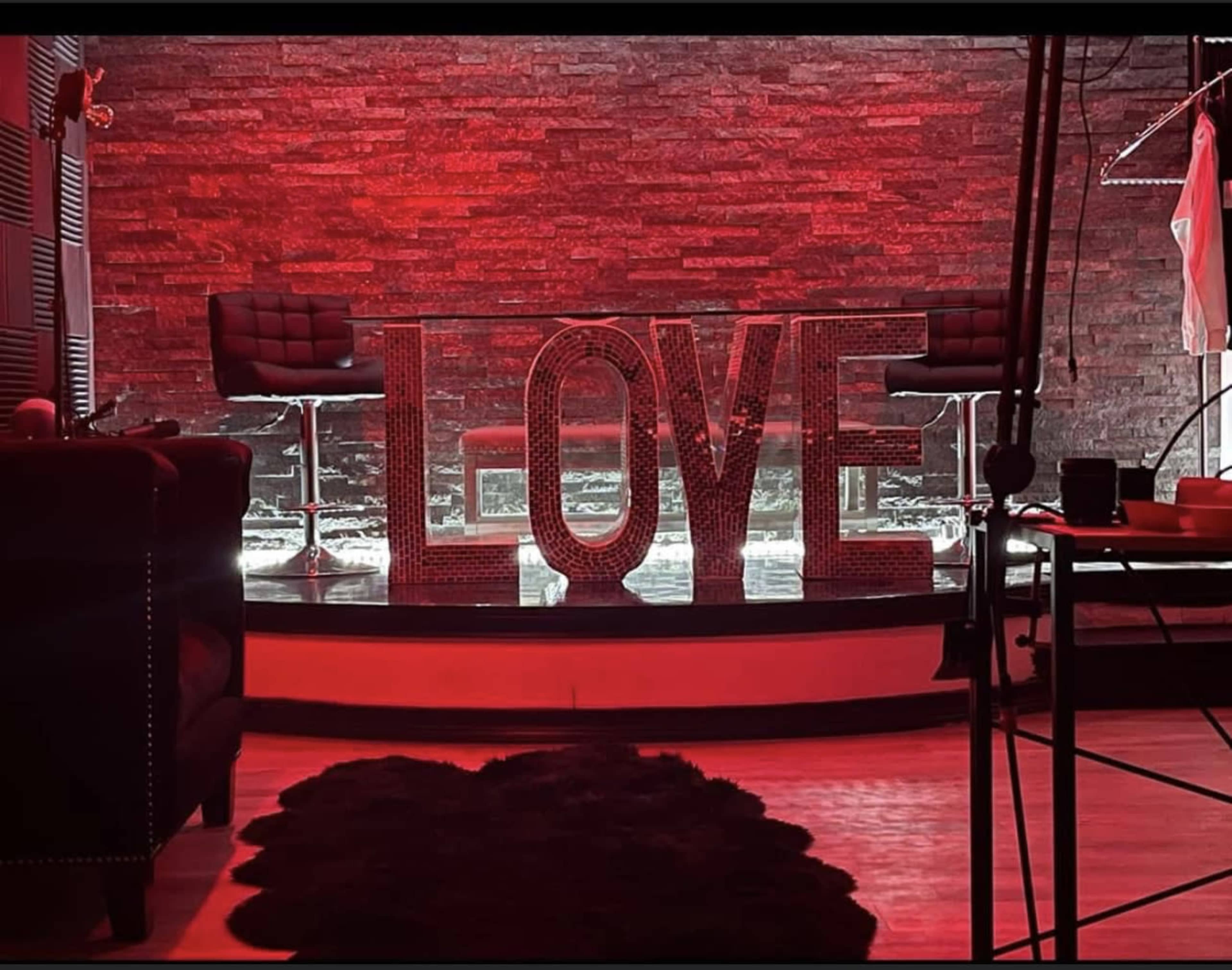 The image features a modern setting with large illuminated letters spelling "LOVE" against a backdrop of red lighting and a stone wall.