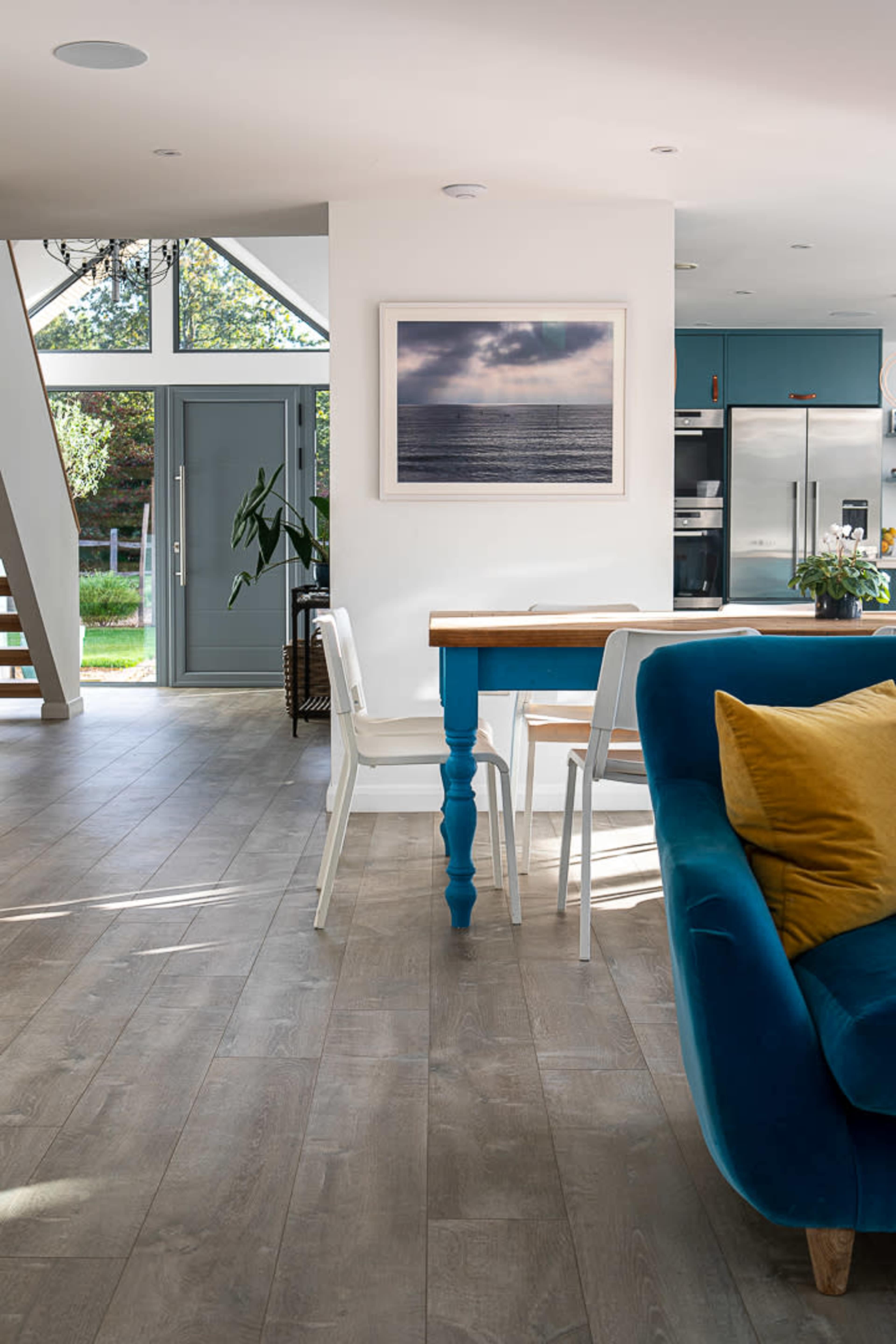 A modern interior space featuring a dining table with blue legs, white chairs, and a large window, set against a backdrop of wooden flooring and a light-colored wall.