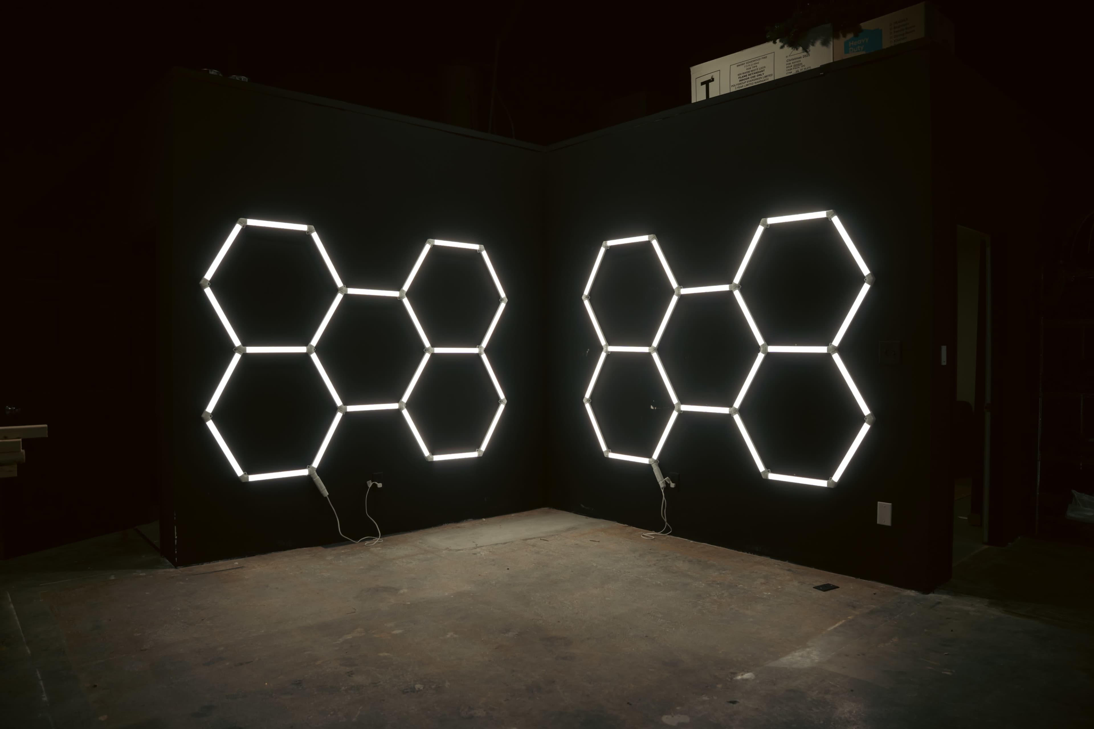 Two large hexagonal light fixtures are mounted on a black wall, creating geometric patterns on either side of the corner in a dimly lit space.