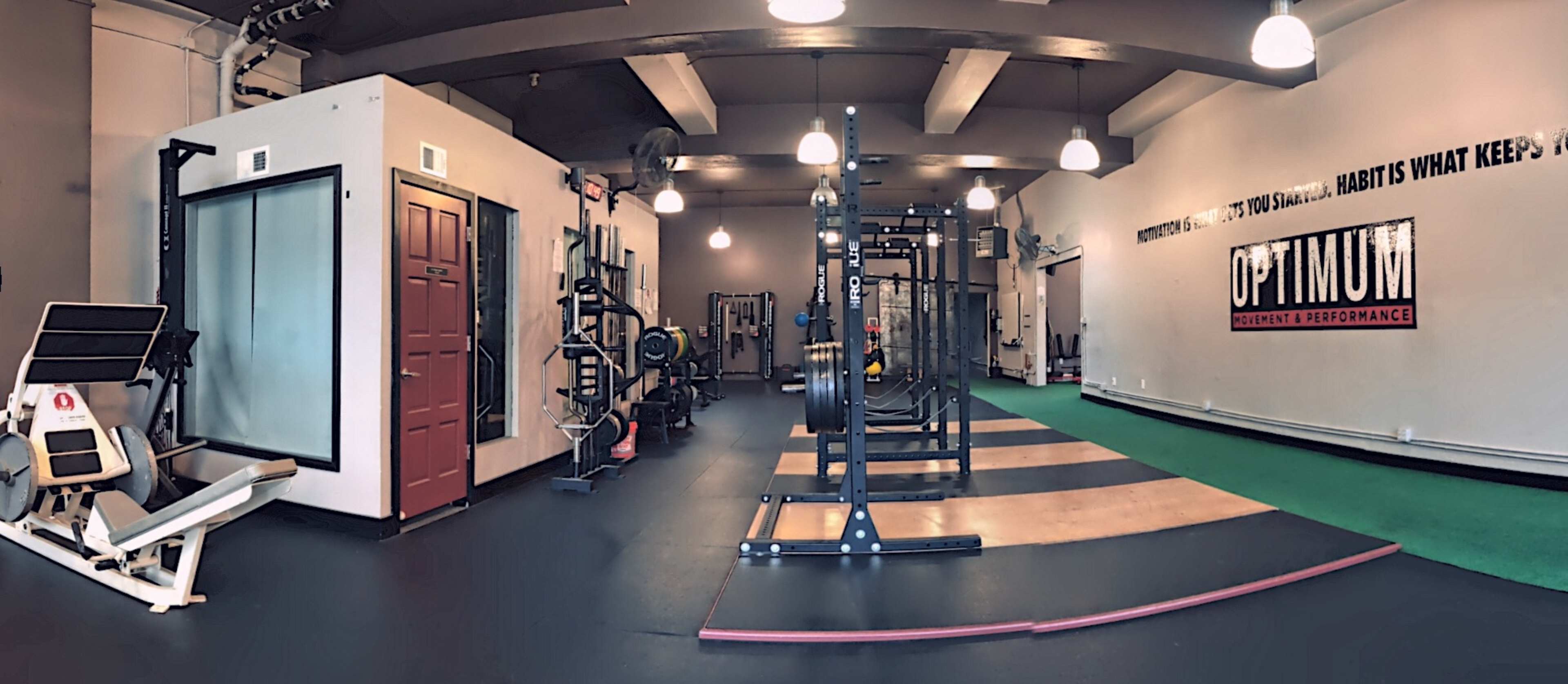 A spacious gym interior with various exercise equipment, including weightlifting racks and cardio machines, arranged along the walls and a green exercise mat in the center.