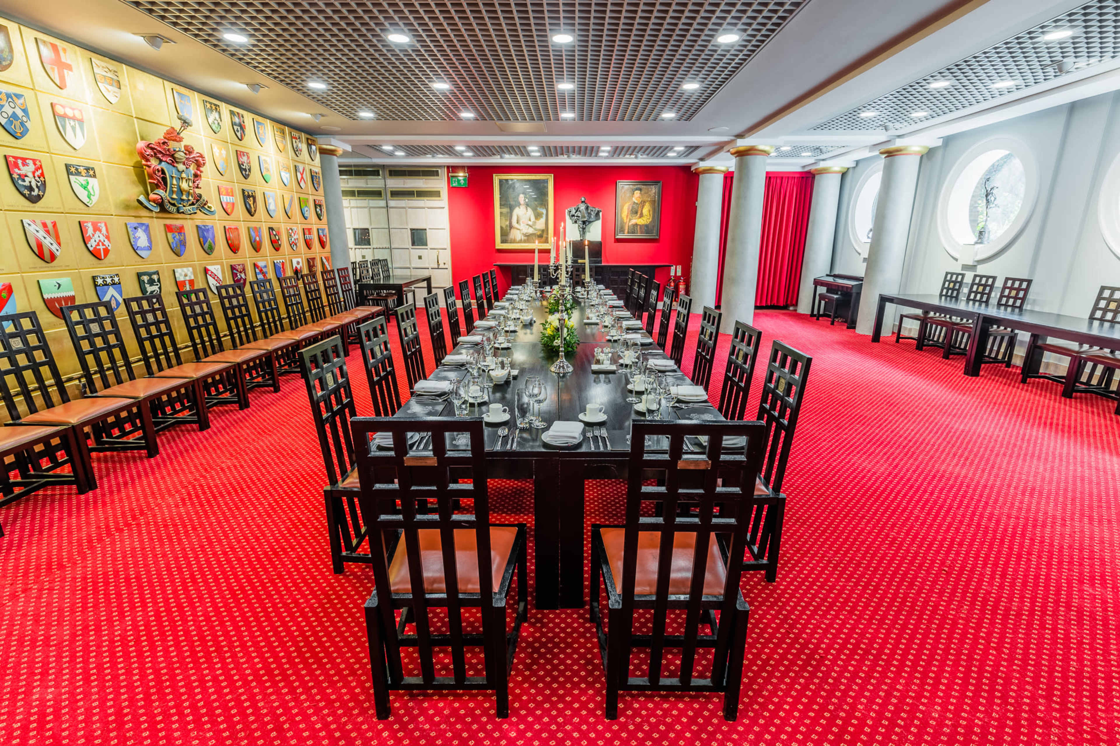 A large, elegantly arranged dining room with a long table set for a formal meal, surrounded by numerous chairs, and adorned with decorative shields on the walls.