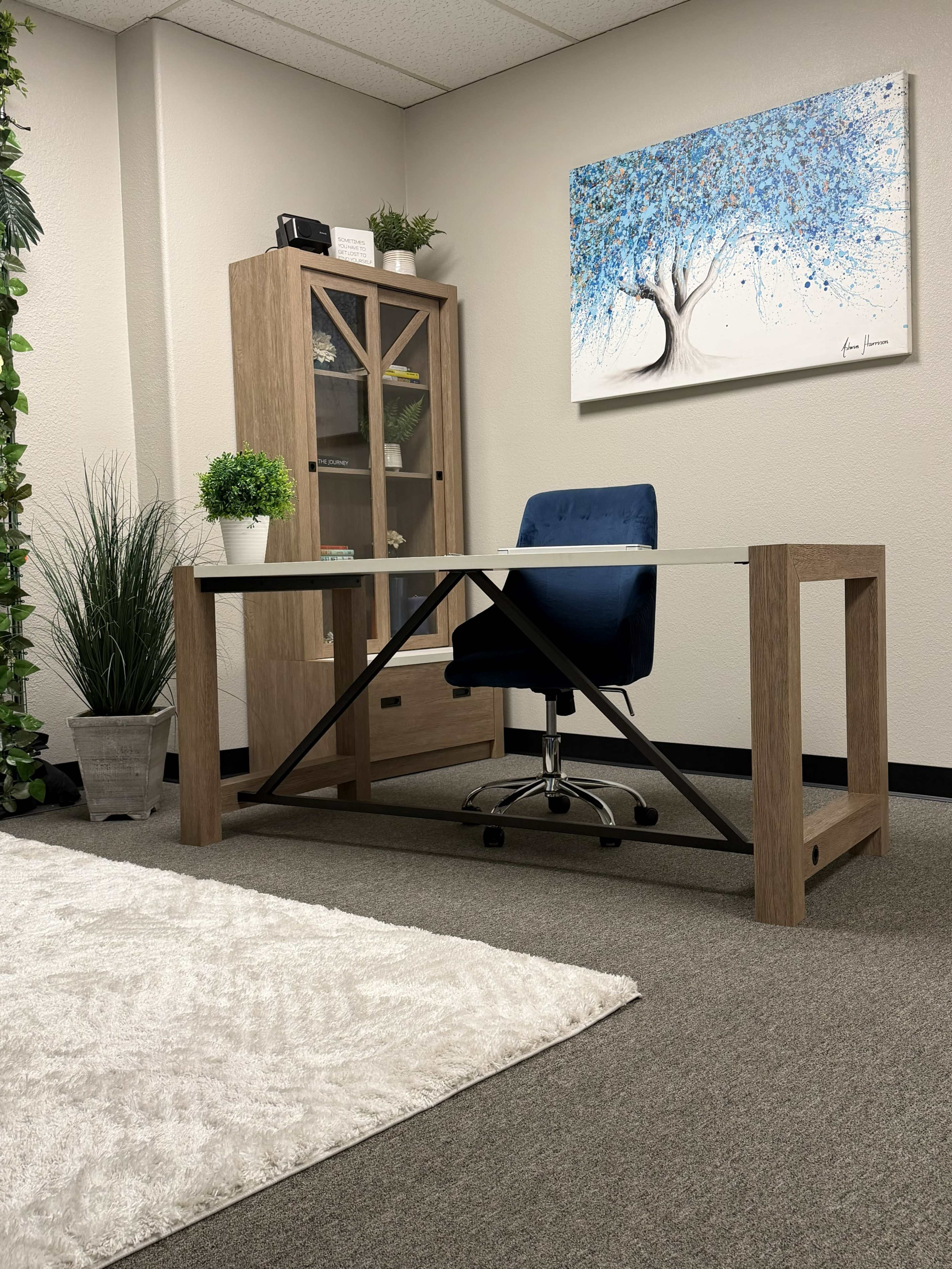 The image shows a modern office space with a glass-top desk, a blue office chair, a wooden cabinet, and a wall art depicting a tree.