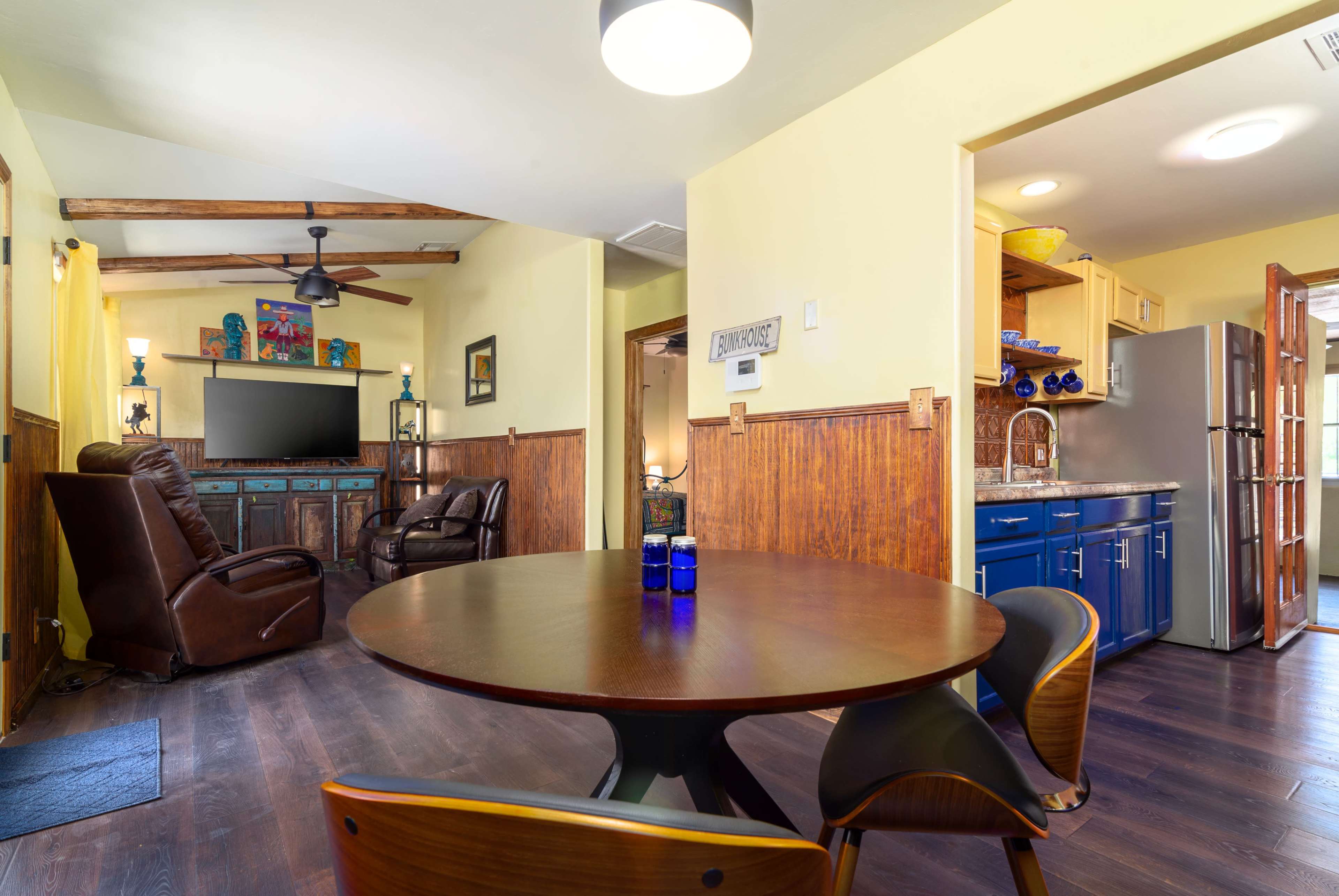 The image shows a cozy living space featuring a round dining table, two brown leather chairs, a green sideboard, and a kitchen area with blue cabinets and modern appliances.