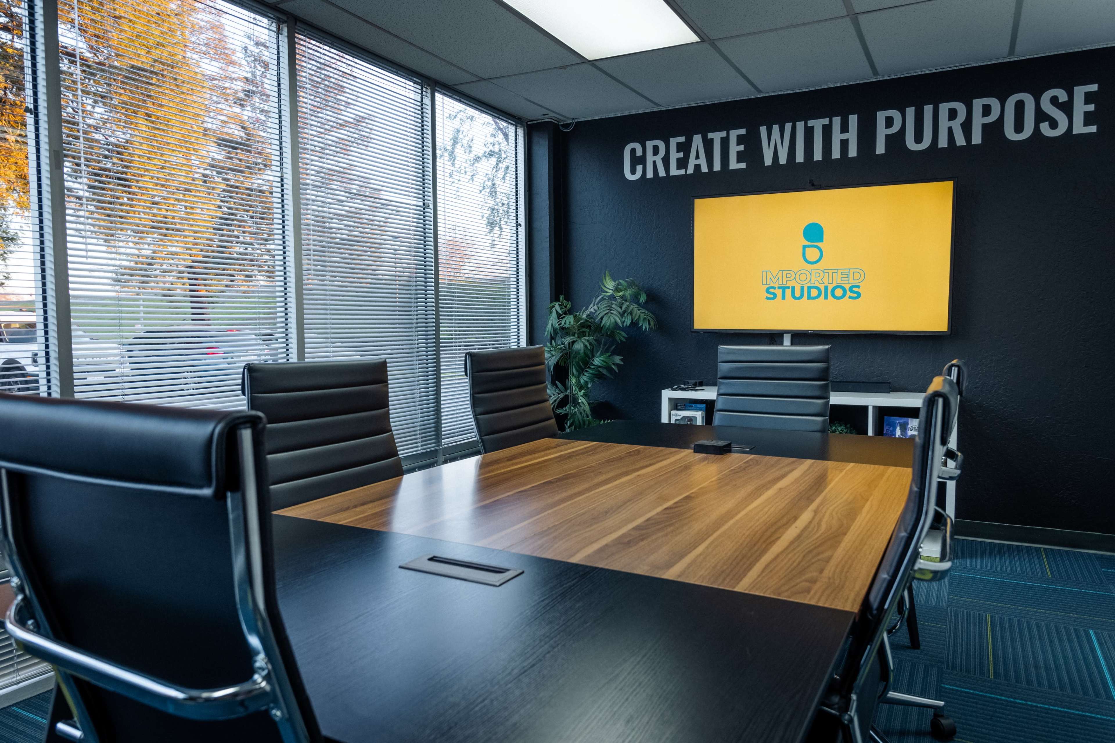 A modern conference room features a large wooden table surrounded by black chairs, with a screen displaying the logo of "Mediated Studios" on the wall.