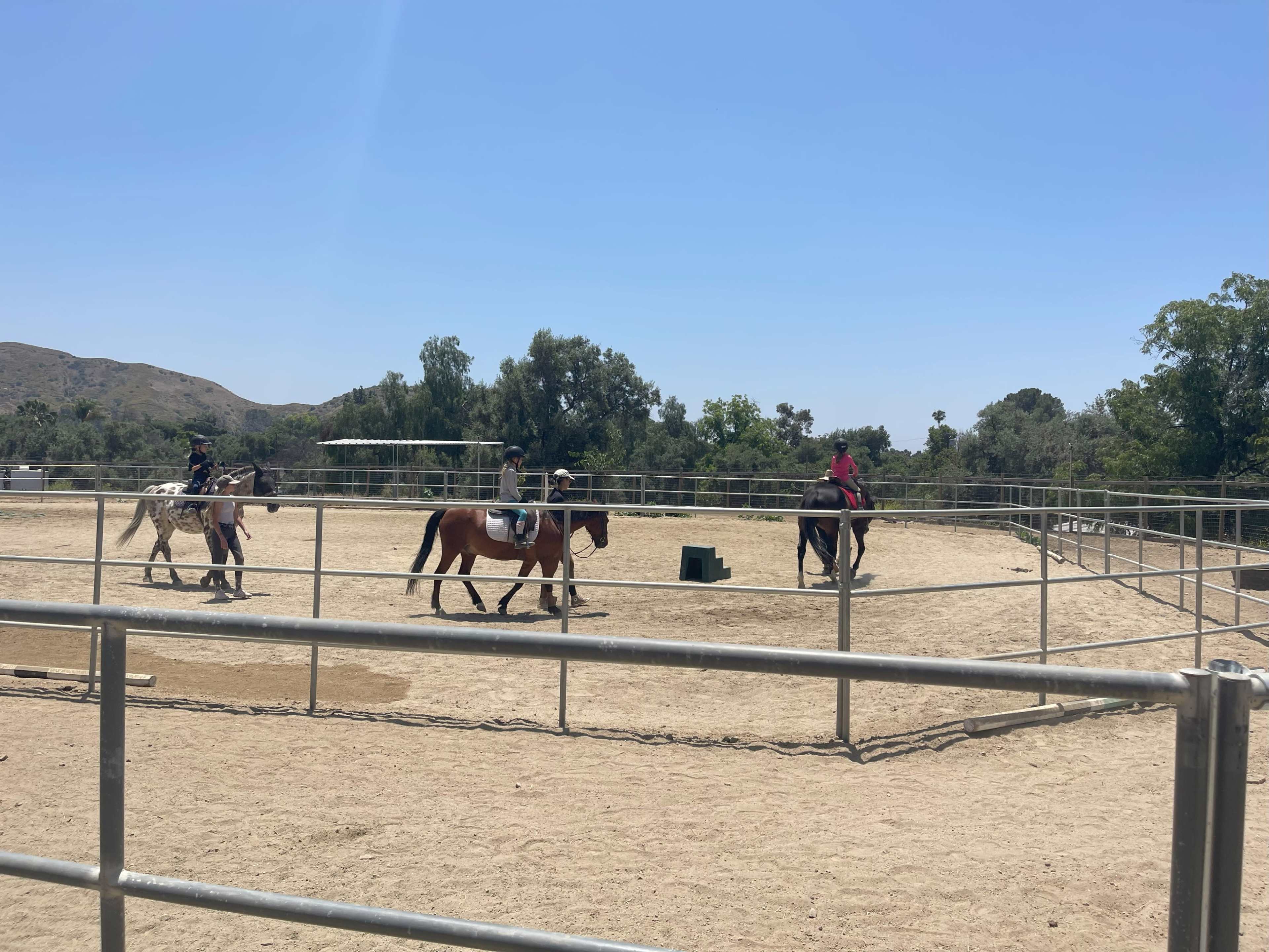 Two riders are on horseback in a sandy arena surrounded by trees and hills under a clear sky.