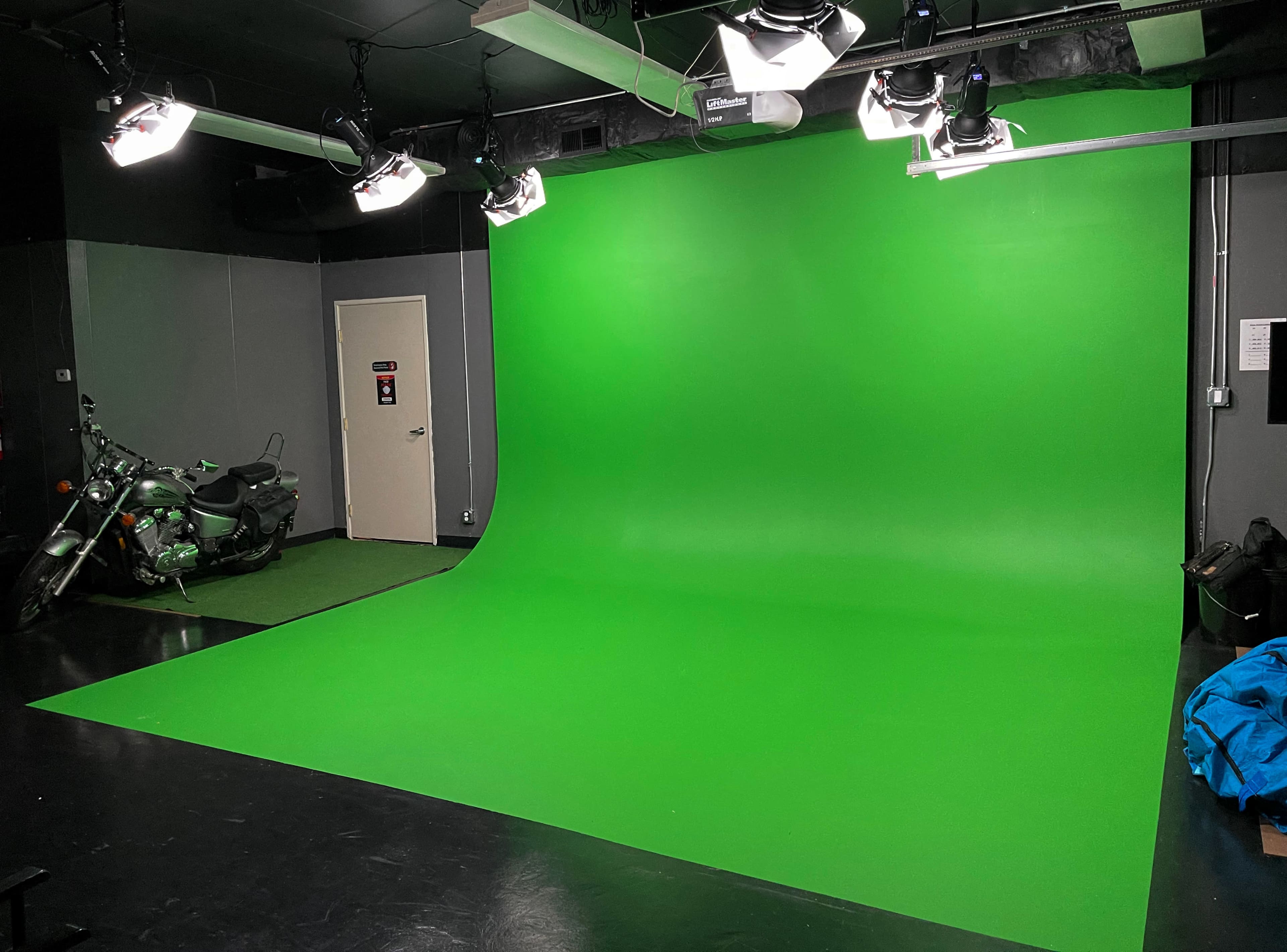 The image shows a studio space featuring a large green screen background, equipped with lighting fixtures and a motorcycle positioned on the floor.