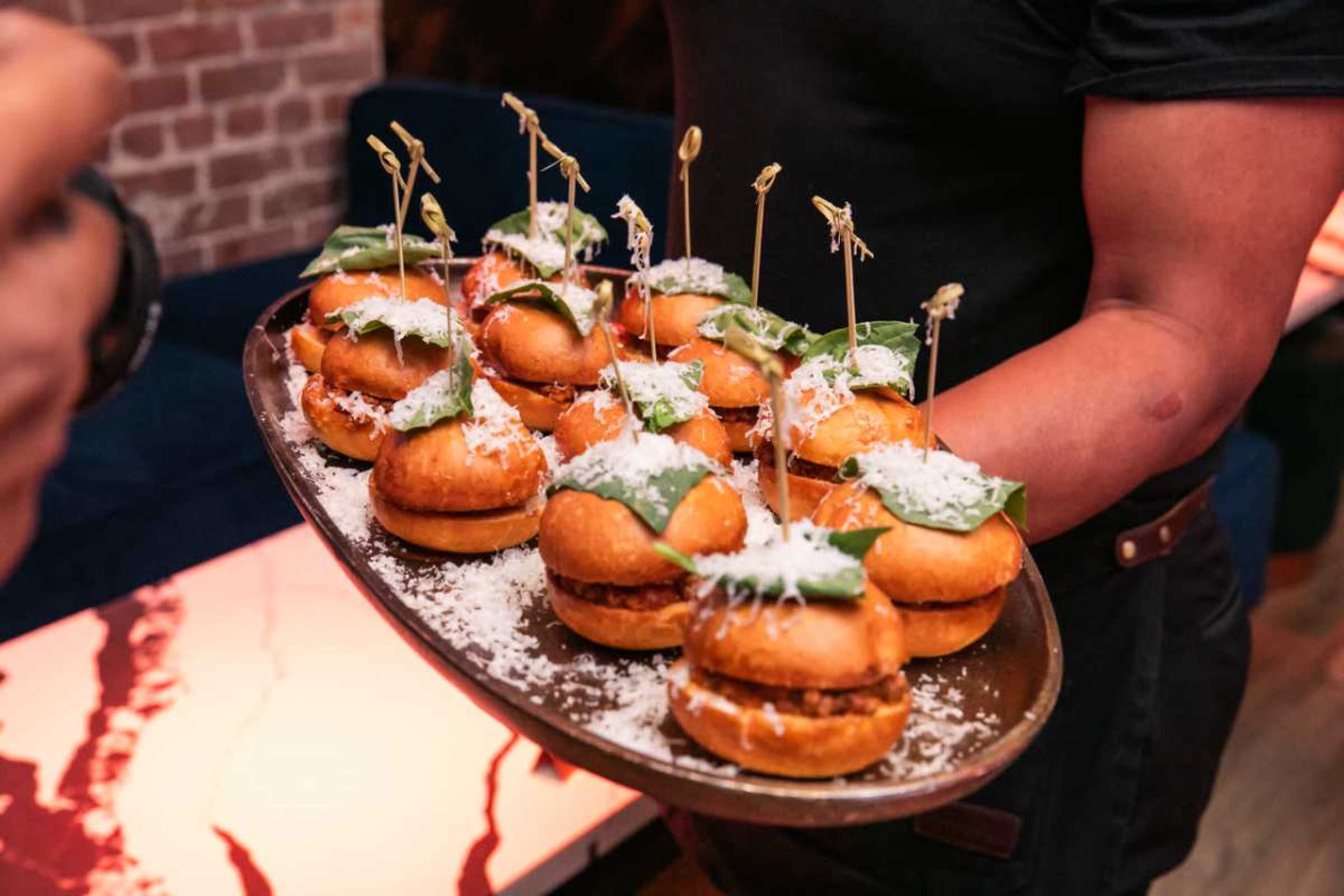 A server holds a tray of small burgers topped with greens and garnished with toothpicks.