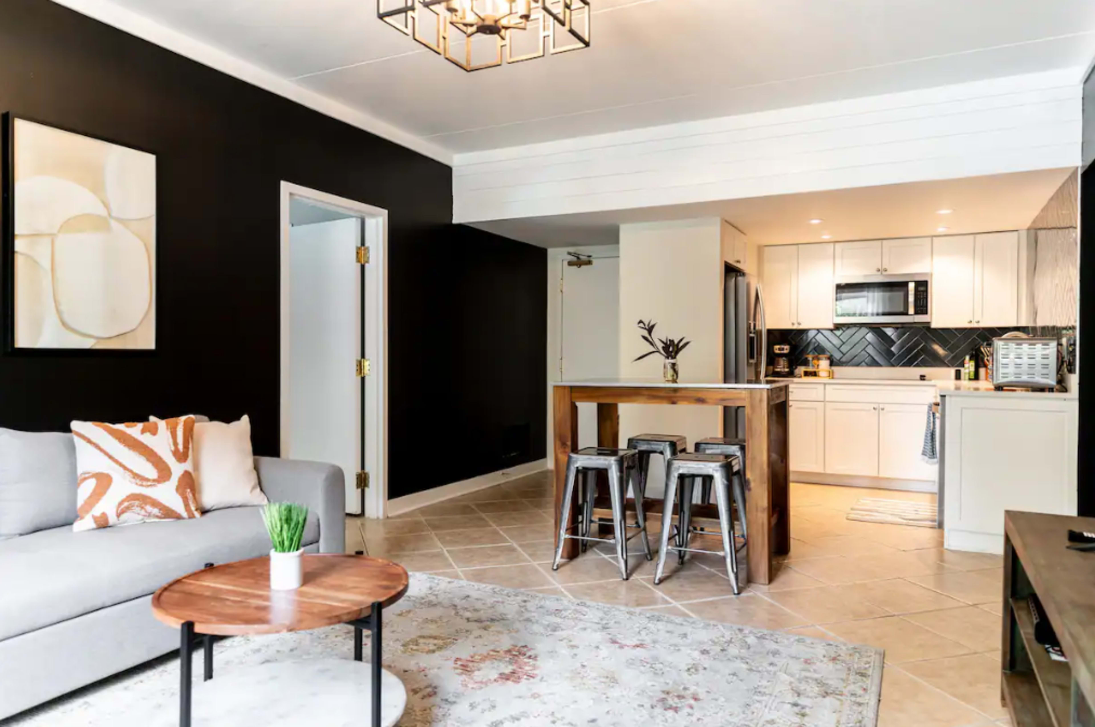 A modern living space with a gray sofa and a round coffee table in front of a kitchen area featuring white cabinets, a microwave, and a tiled backsplash.
