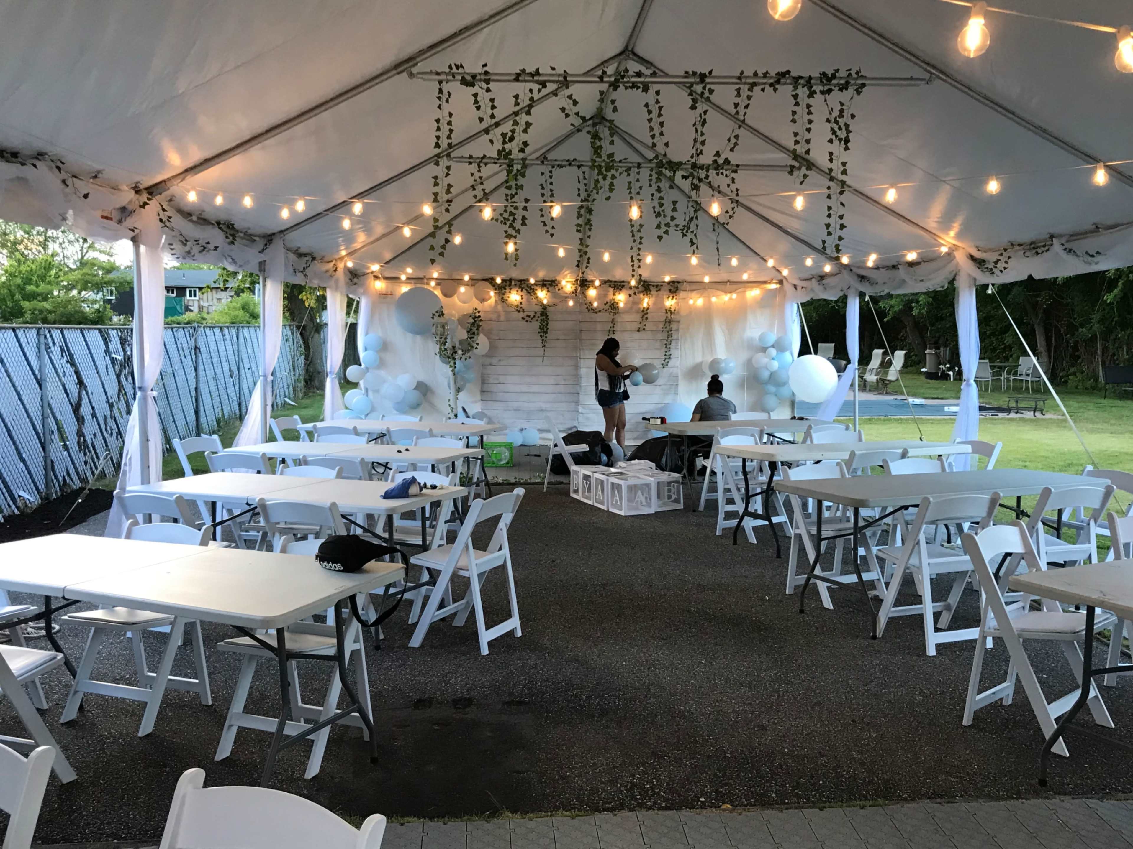 A tented event space features white tables and chairs arranged under string lights and decorative greenery.