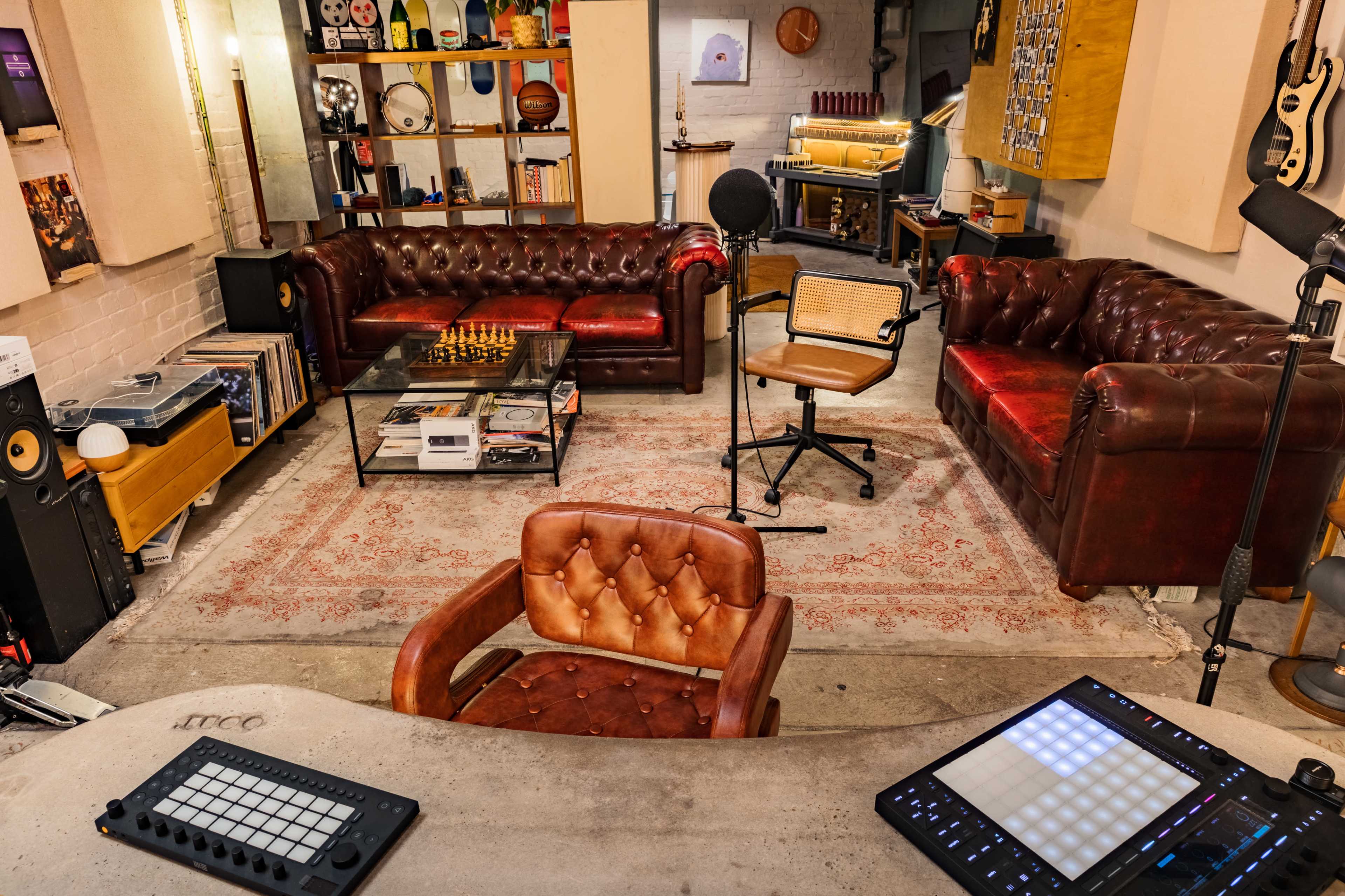 The image shows a cozy music studio with leather couches, a glass coffee table, a microphone, and a chair at a desk featuring electronic equipment.