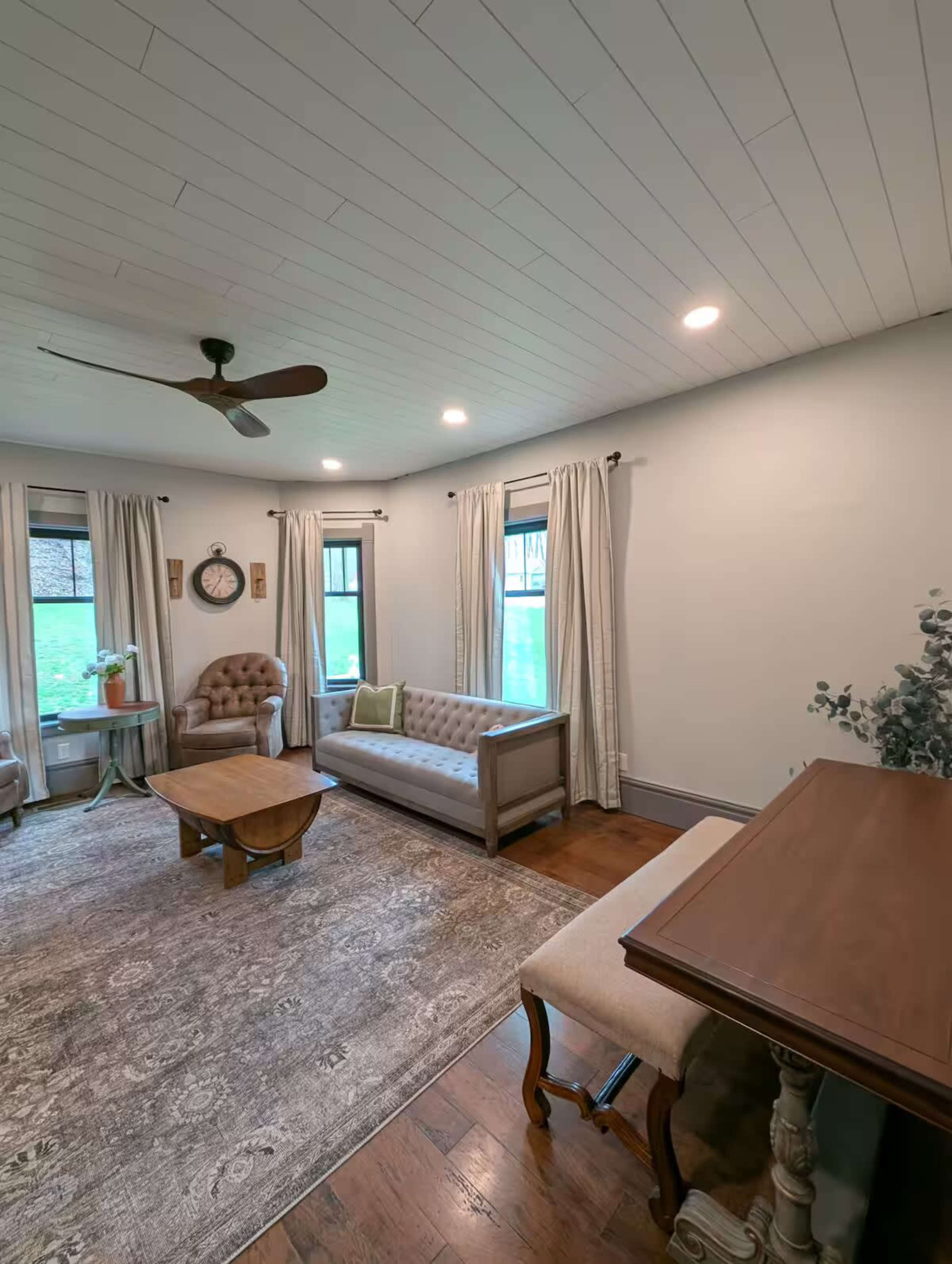 The image shows a living room furnished with a beige sofa, a wooden coffee table, a tufted chair, and natural light coming through two windows.