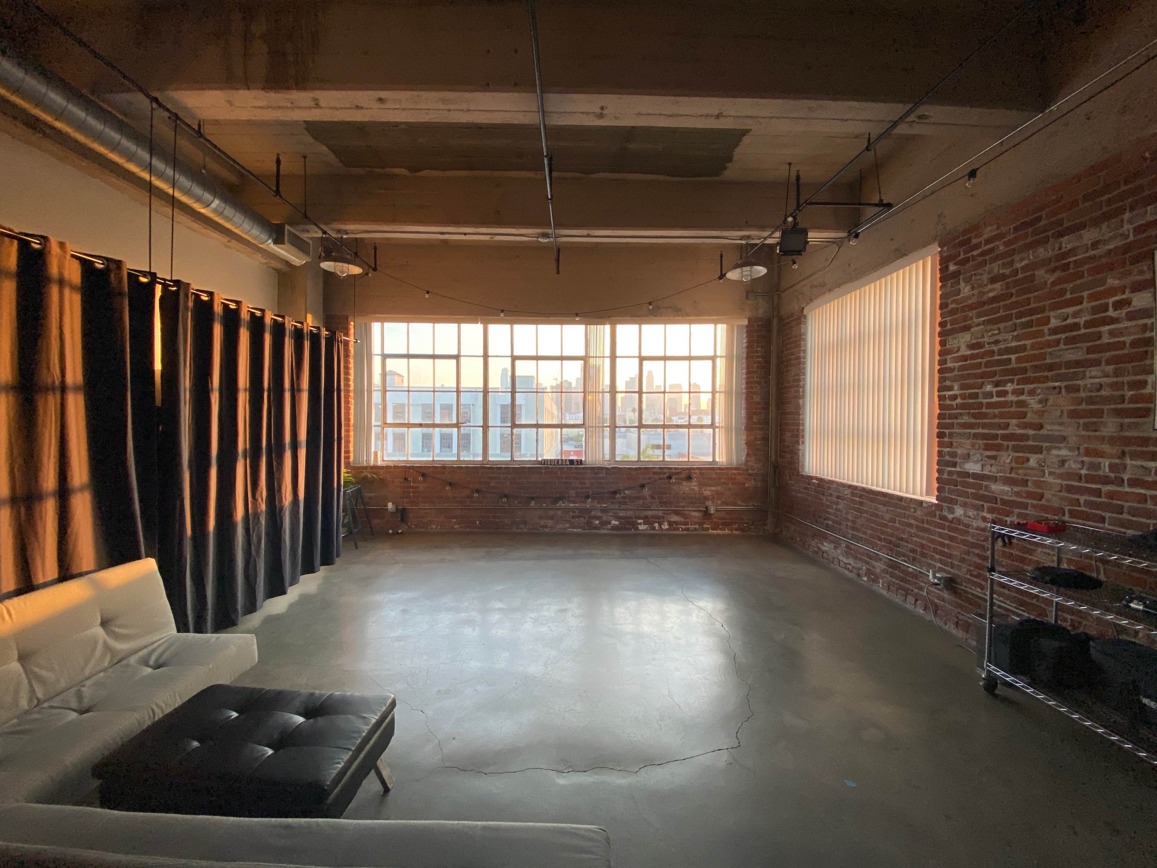 A spacious room features large windows with sunlight streaming in, brick walls, and minimal furniture including a white couch and a coffee table.