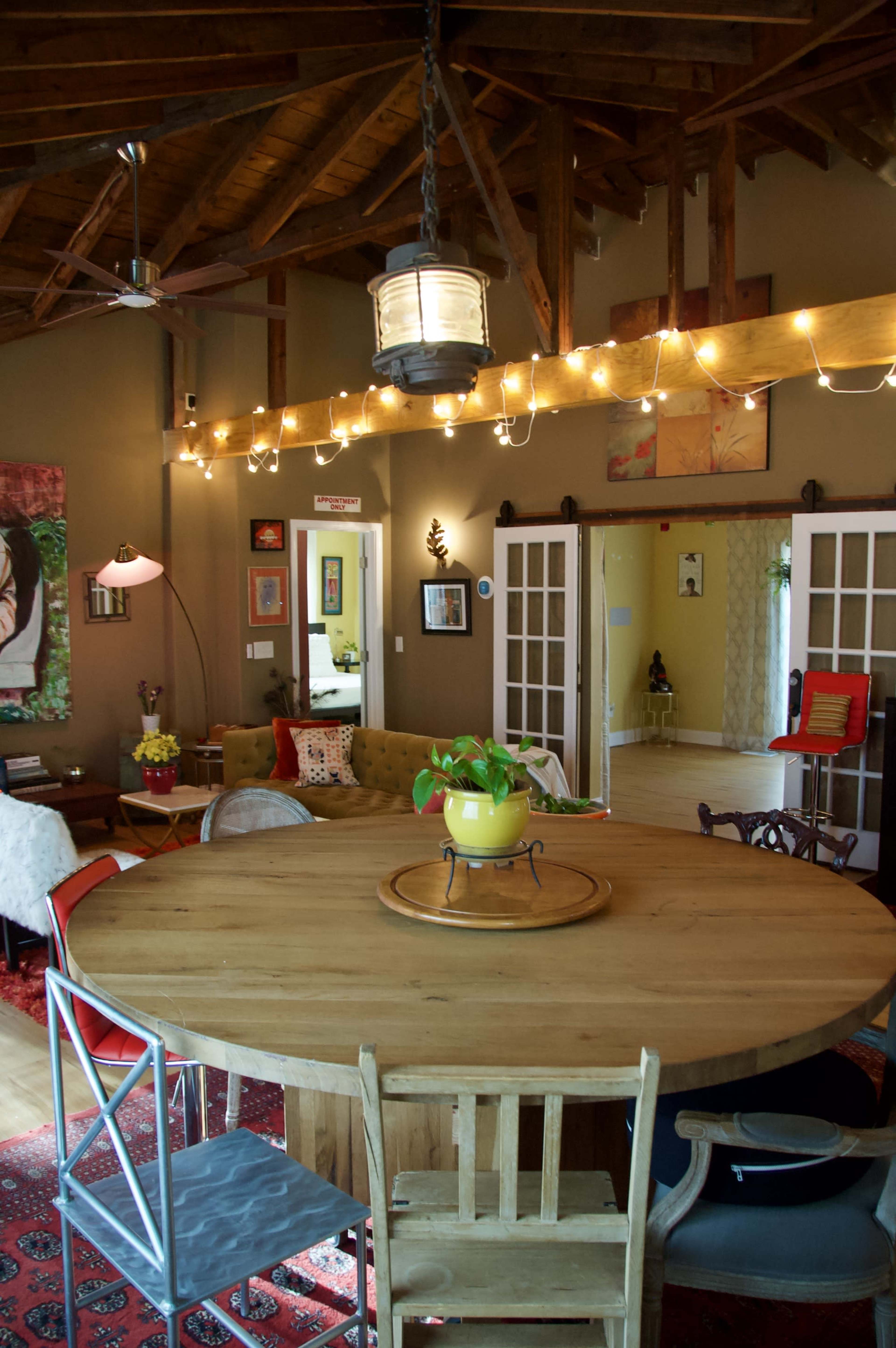 A round wooden table is centered in a spacious, well-lit room with exposed beams and string lights, surrounded by various chairs and colorful decor.