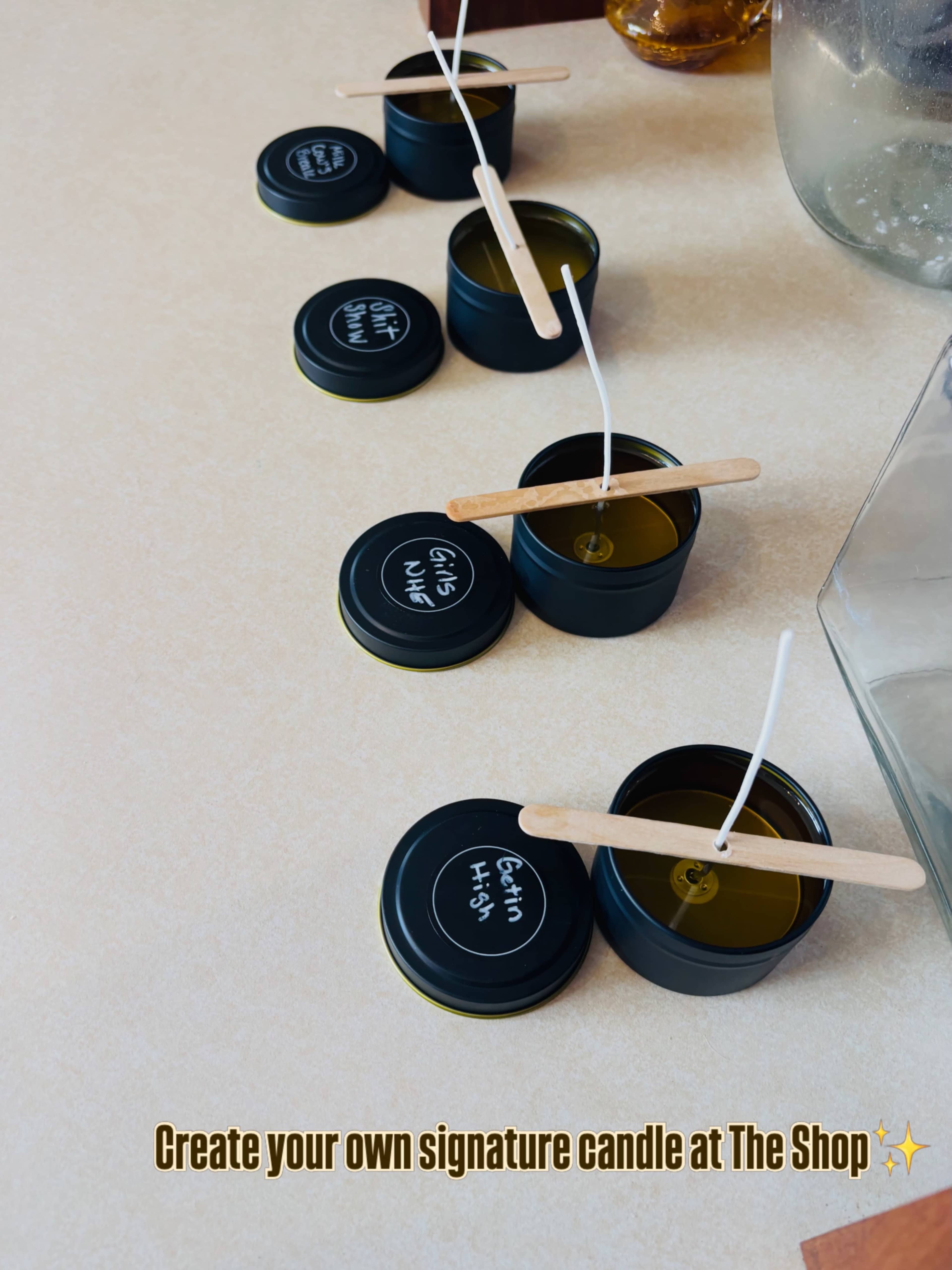 The image shows several black candle containers with wooden stirring sticks and candle wicks, arranged on a countertop, with a sign inviting people to create their own signature candles.