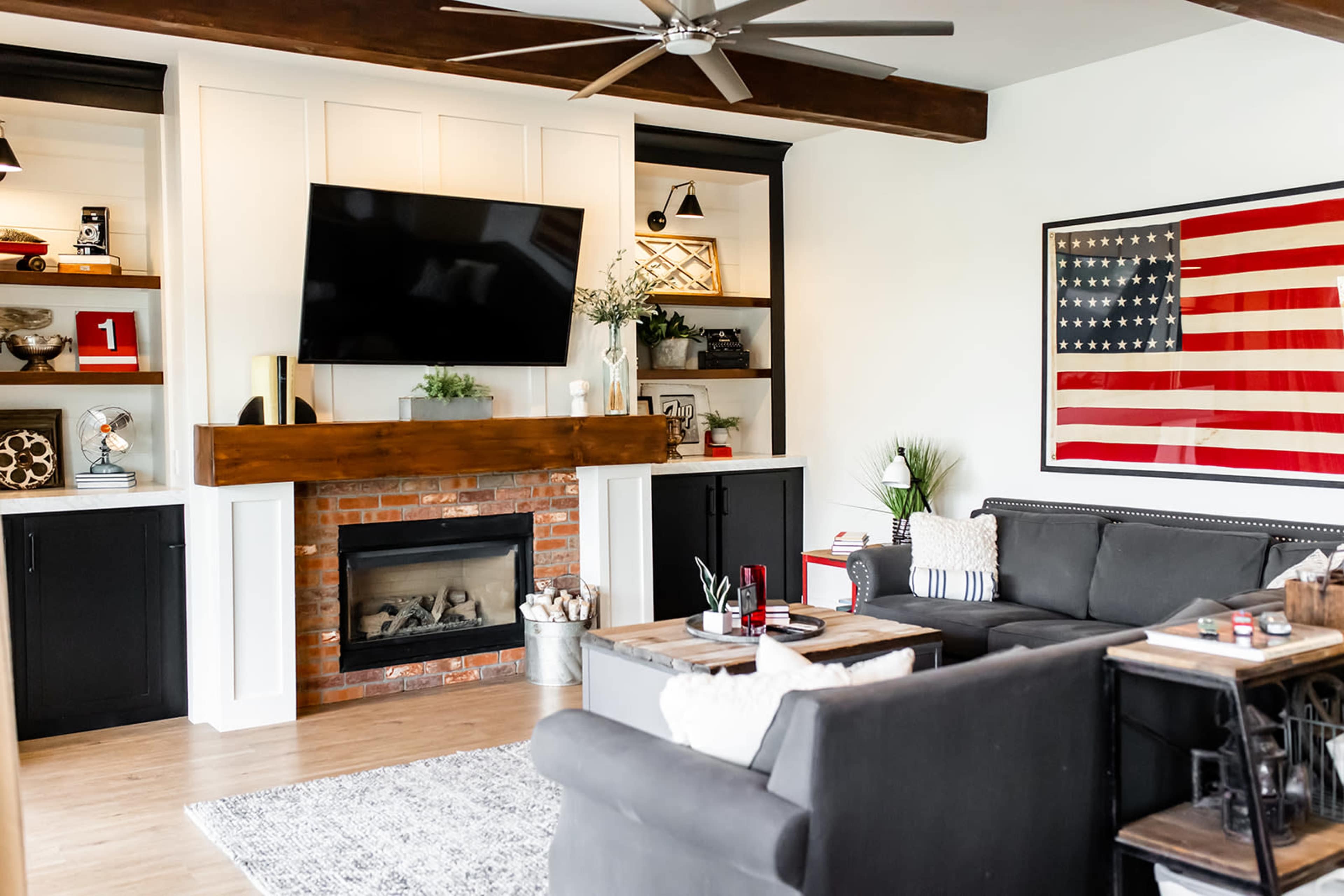 A modern living room features a large television above a brick fireplace, flanked by built-in shelves, with a rustic wooden mantel and an American flag artwork on the wall.