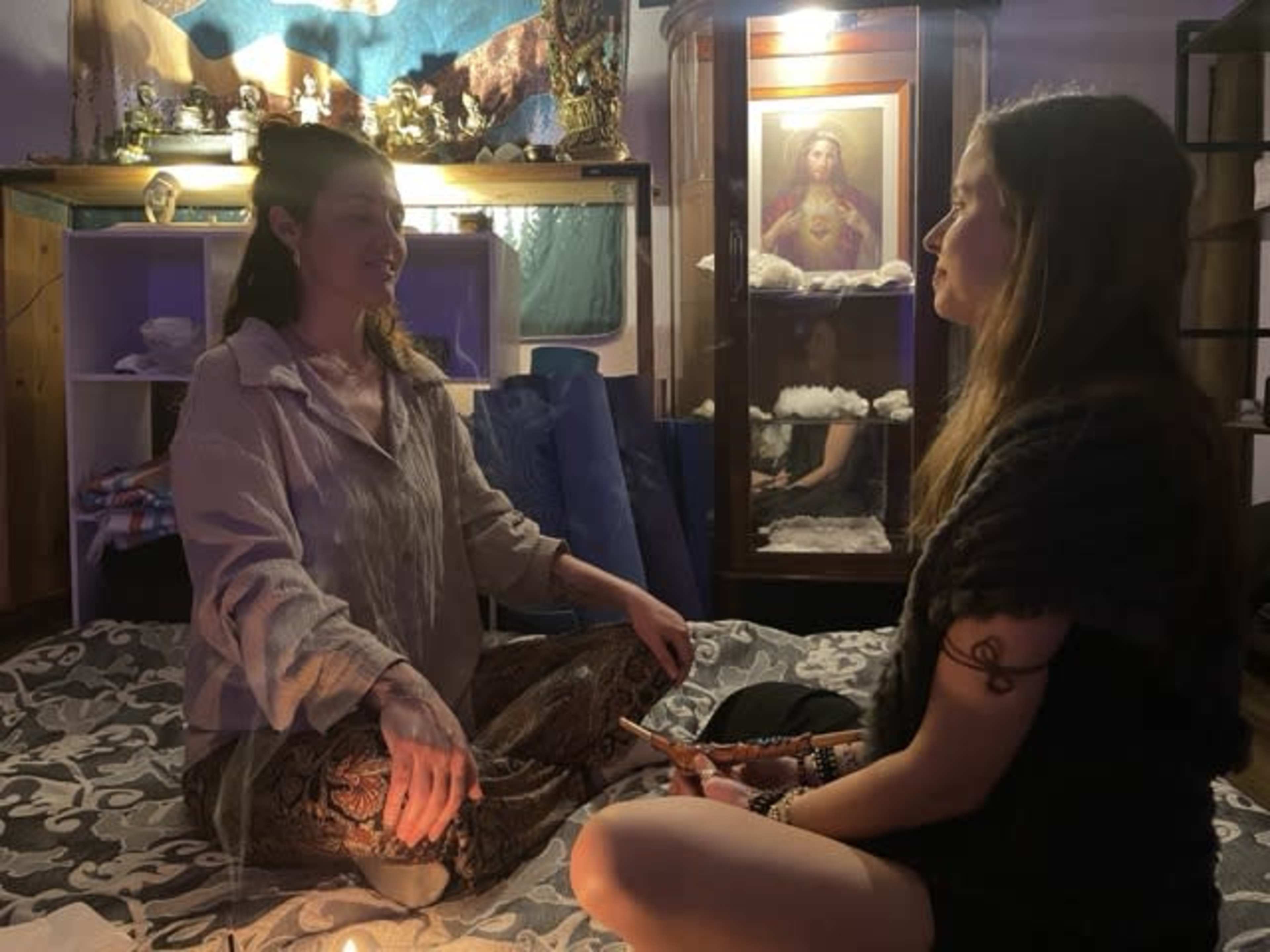 Two women sit cross-legged on a patterned floor, engaged in a focused conversation in a warmly lit room filled with spiritual artifacts.