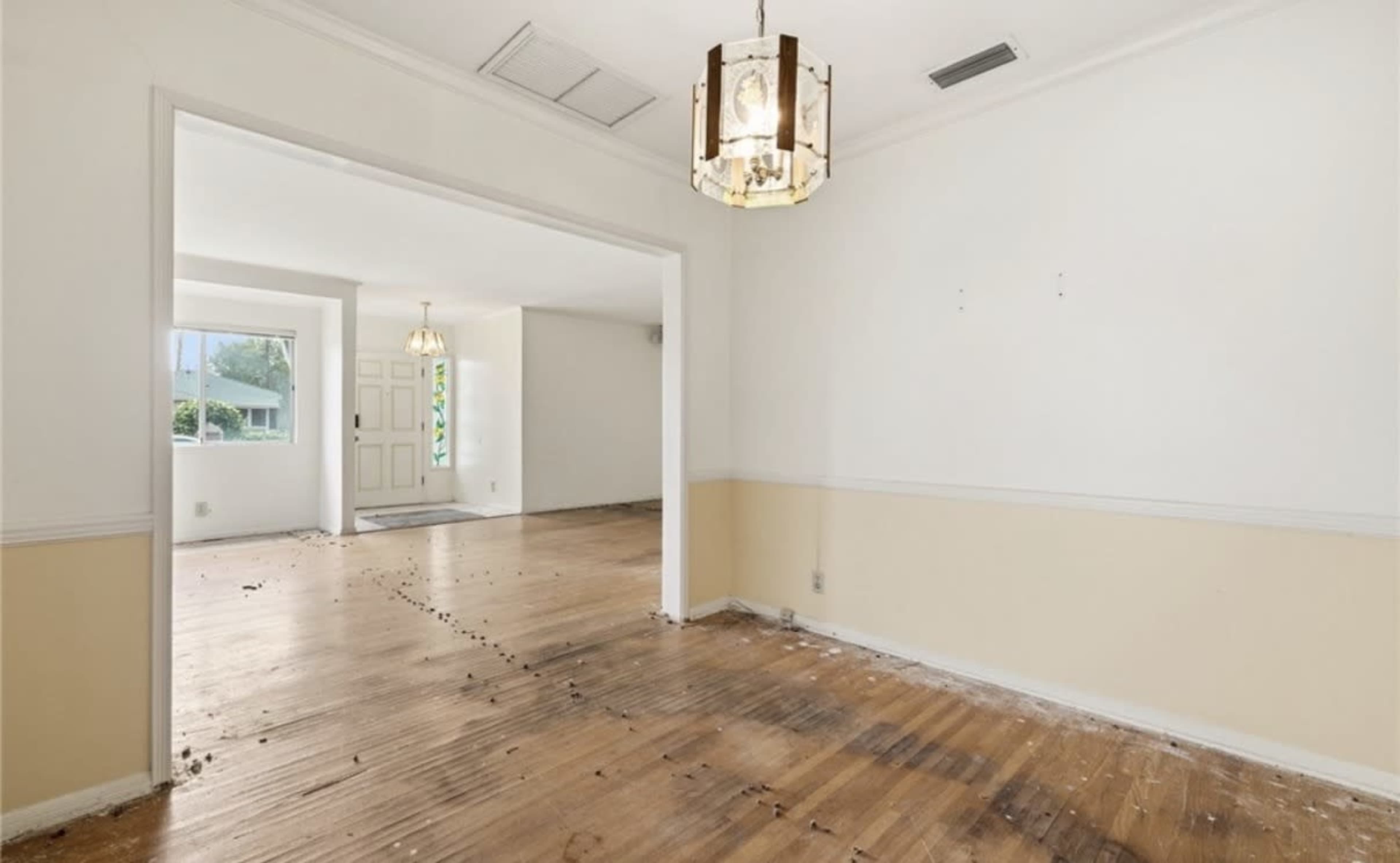 The image shows a partially empty room with wooden floors, a light fixture hanging from the ceiling, and visible signs of wear and debris on the floor.