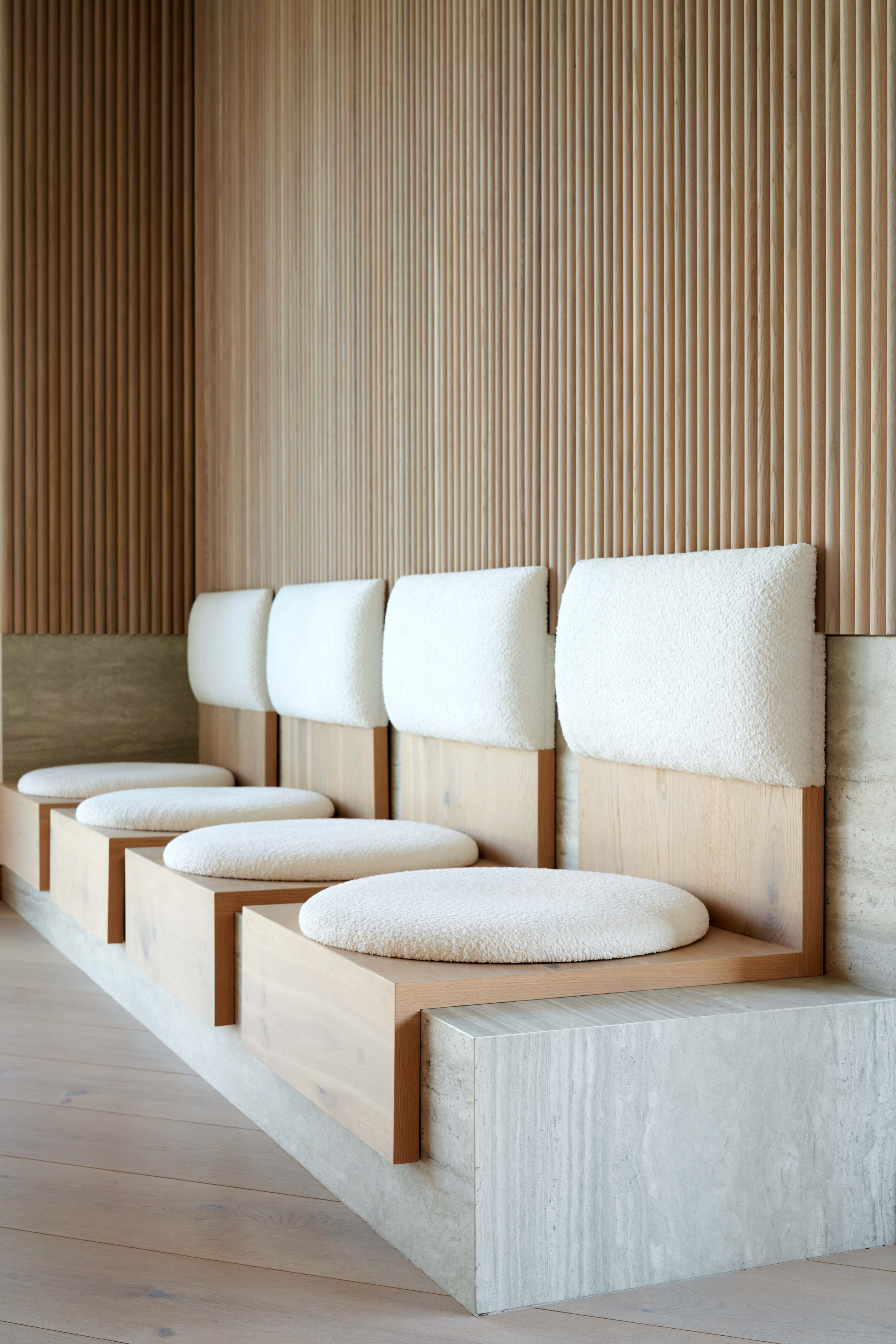 The image shows a modern seating area with a series of wooden benches featuring upholstered backrests and round cushions against a textured wood-paneled wall.
