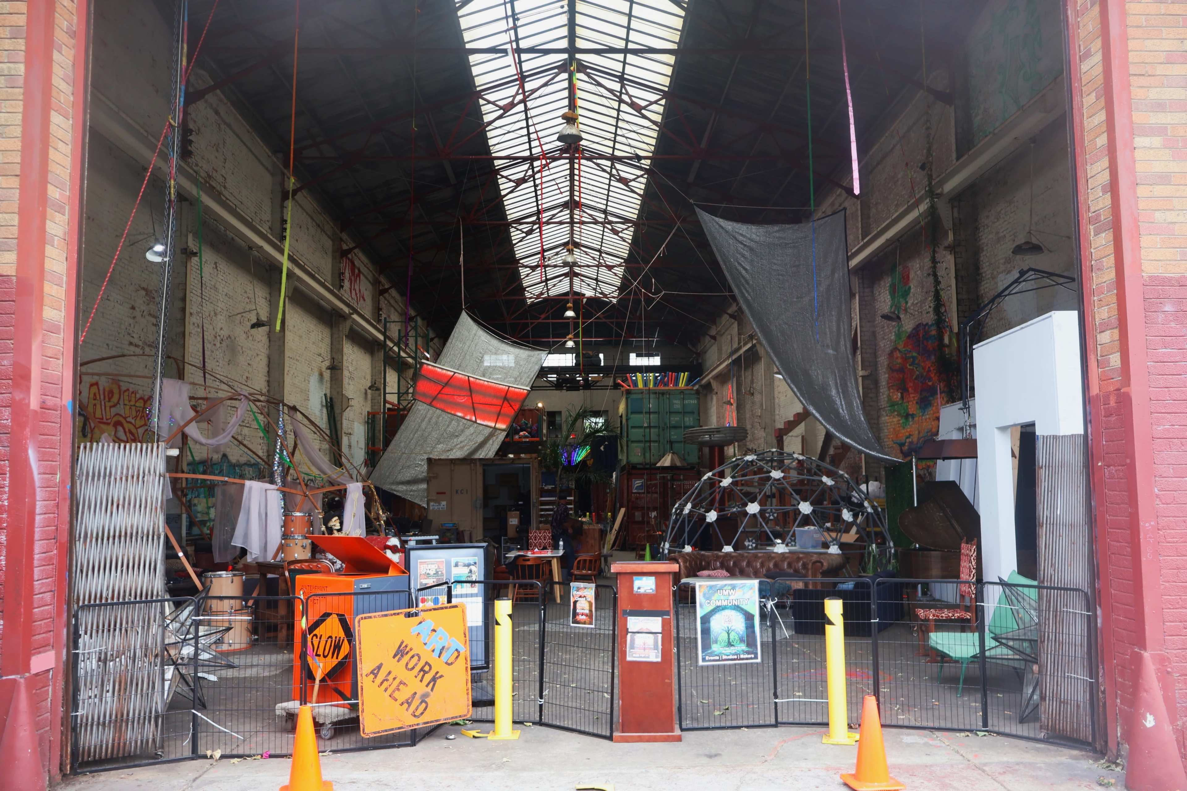 The image shows an industrial warehouse interior filled with various artworks, furniture, and eclectic decorations, with a large open space and makeshift barriers at the entrance.