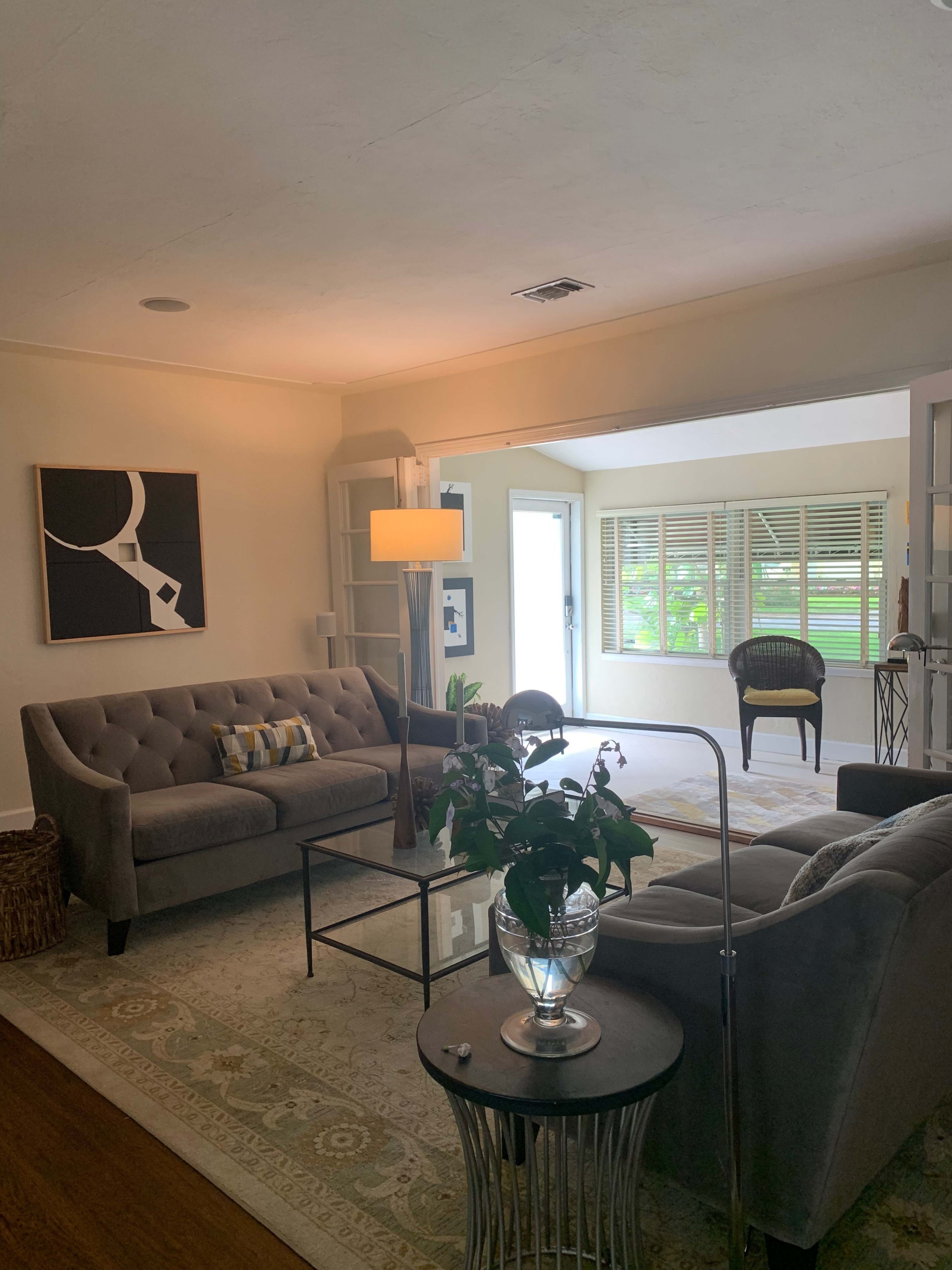 The image shows a living room with gray sofas, a floor lamp, a coffee table, and a large window leading to an outdoor view.