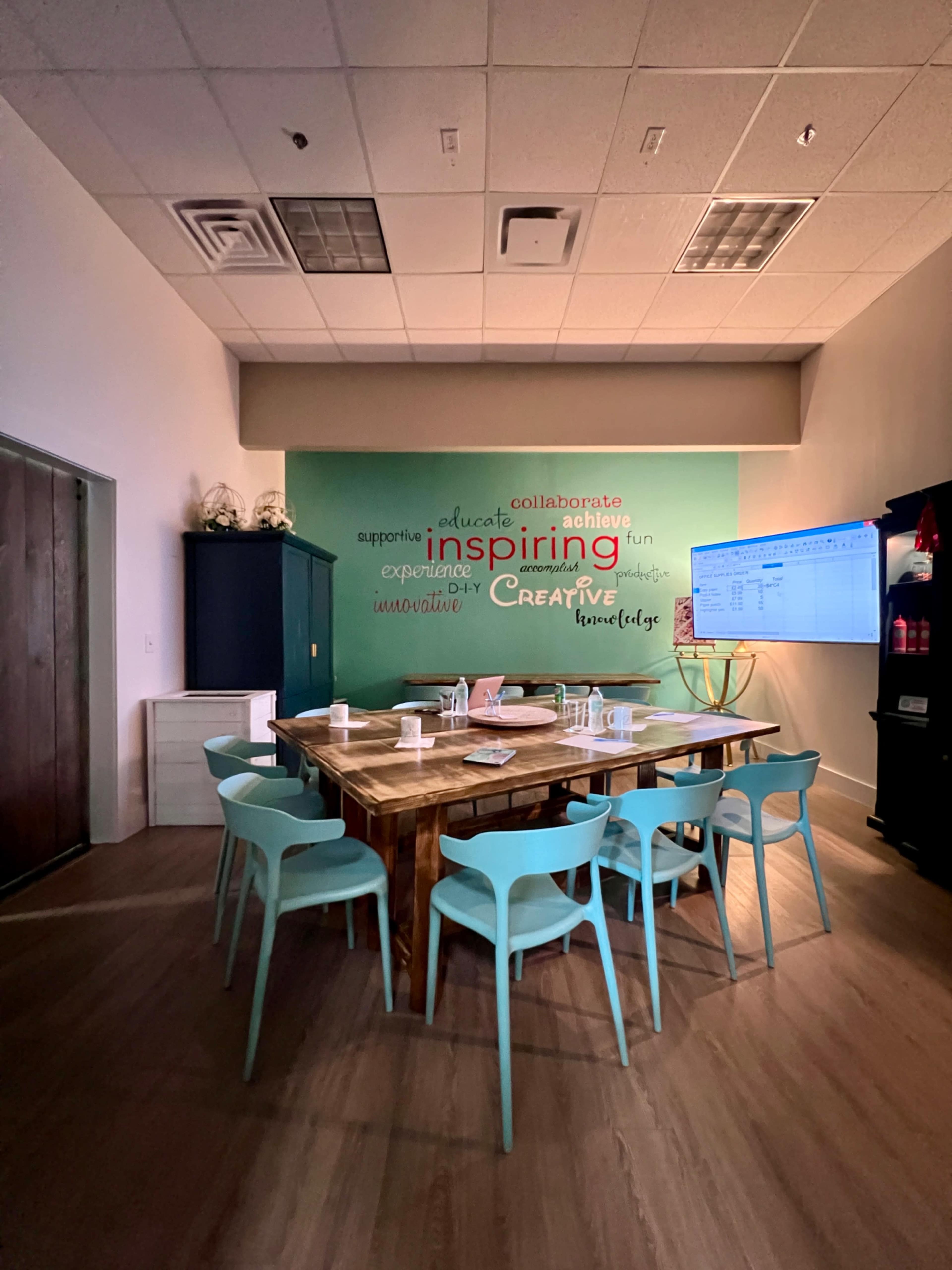 A meeting room features a large wooden table surrounded by light blue chairs, with a green wall displaying motivational words and a screen on one side.
