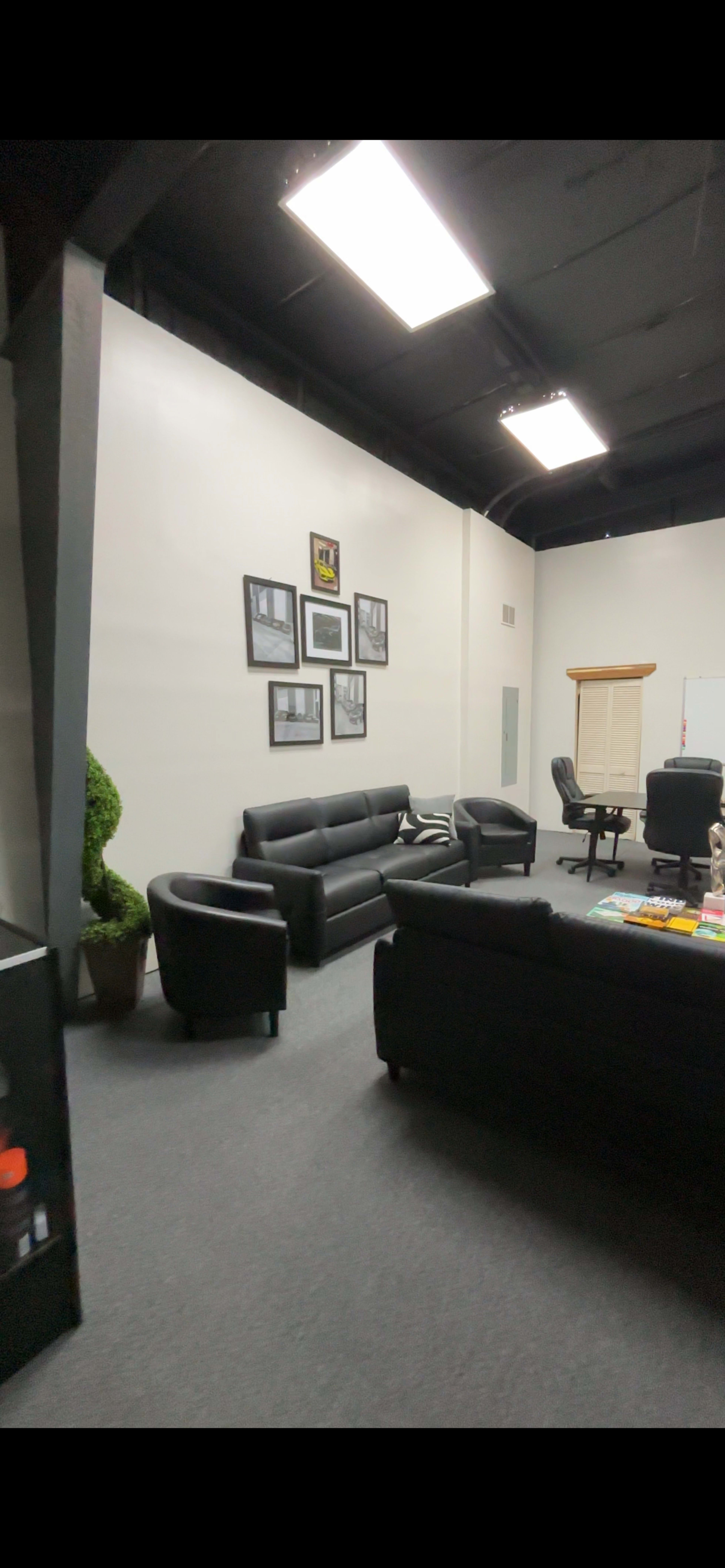 The image shows a spacious office area featuring black couches, a conference chair, framed photographs on the wall, and a potted plant.