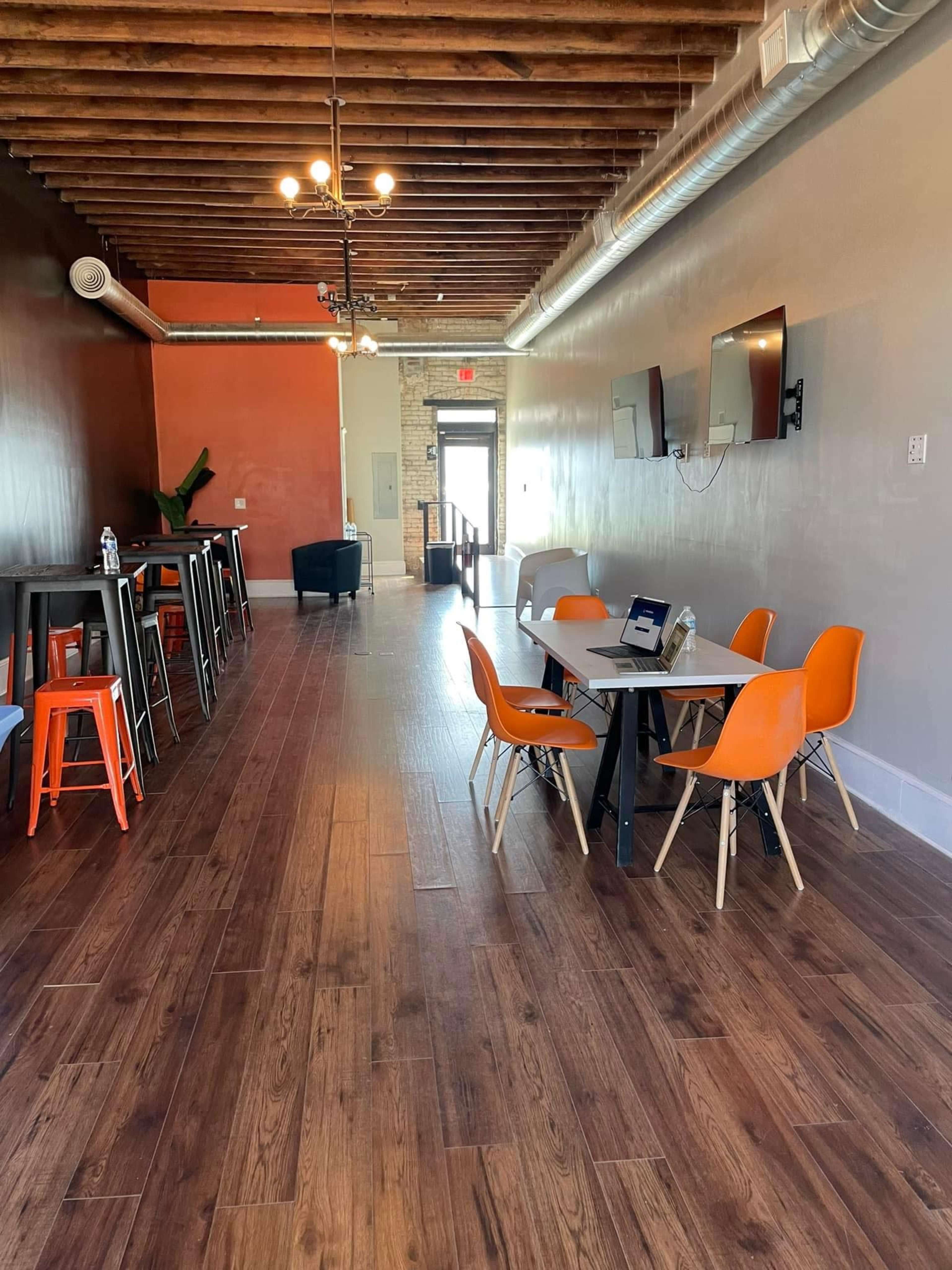 A modern, open interior space featuring wooden floors, a long table with orange and white chairs, a bar area with high stools, and two wall-mounted televisions.